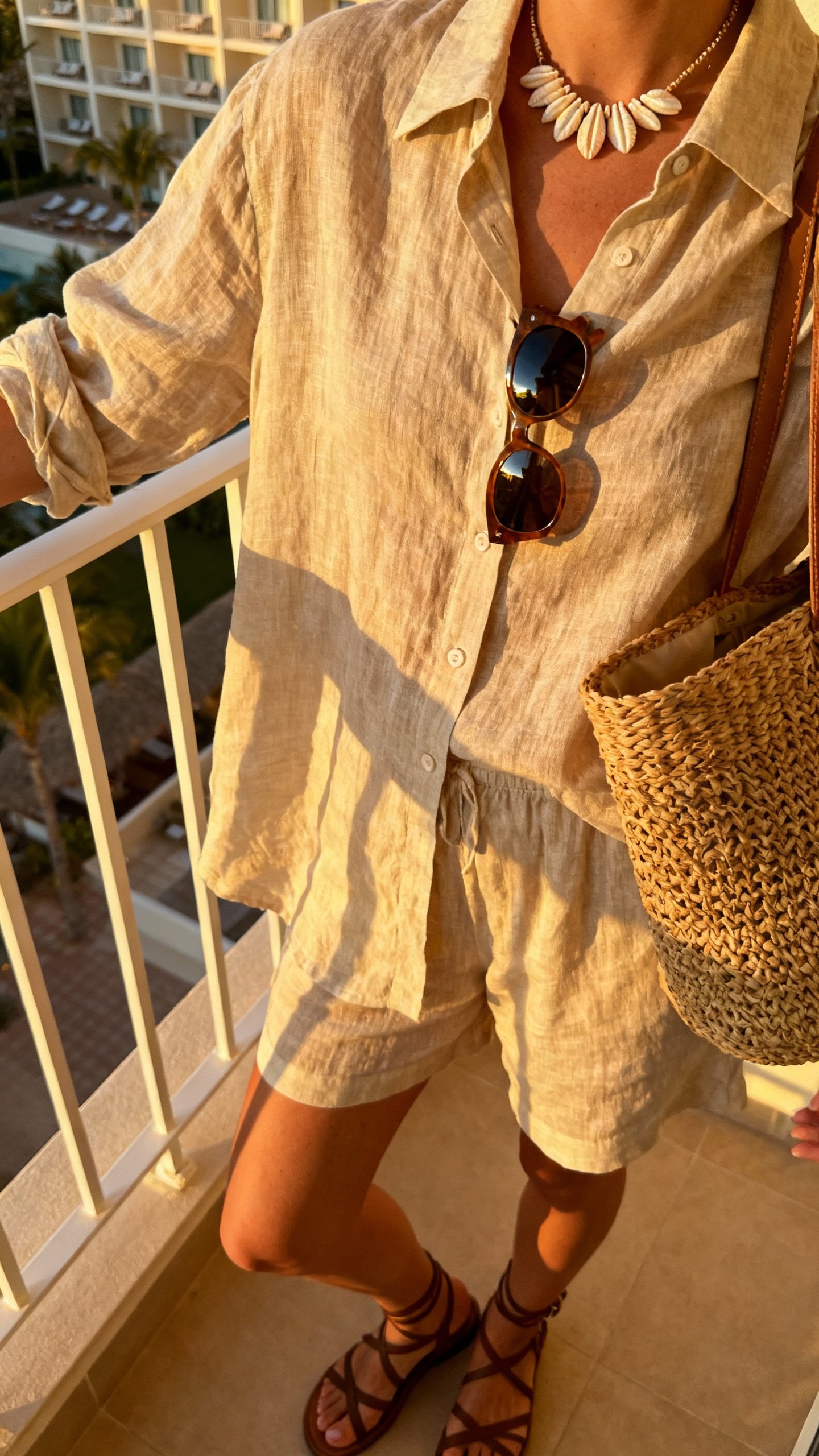 iPhone balcony mirror selfie of a woman in a luxe linen-blend set: relaxed linen-blend button-up and matching shorts, strappy sandals, woven tote, oversized sunglasses, shell necklace, face not visible, travel hotel room balcony with afternoon light, natural iPhone photo quality