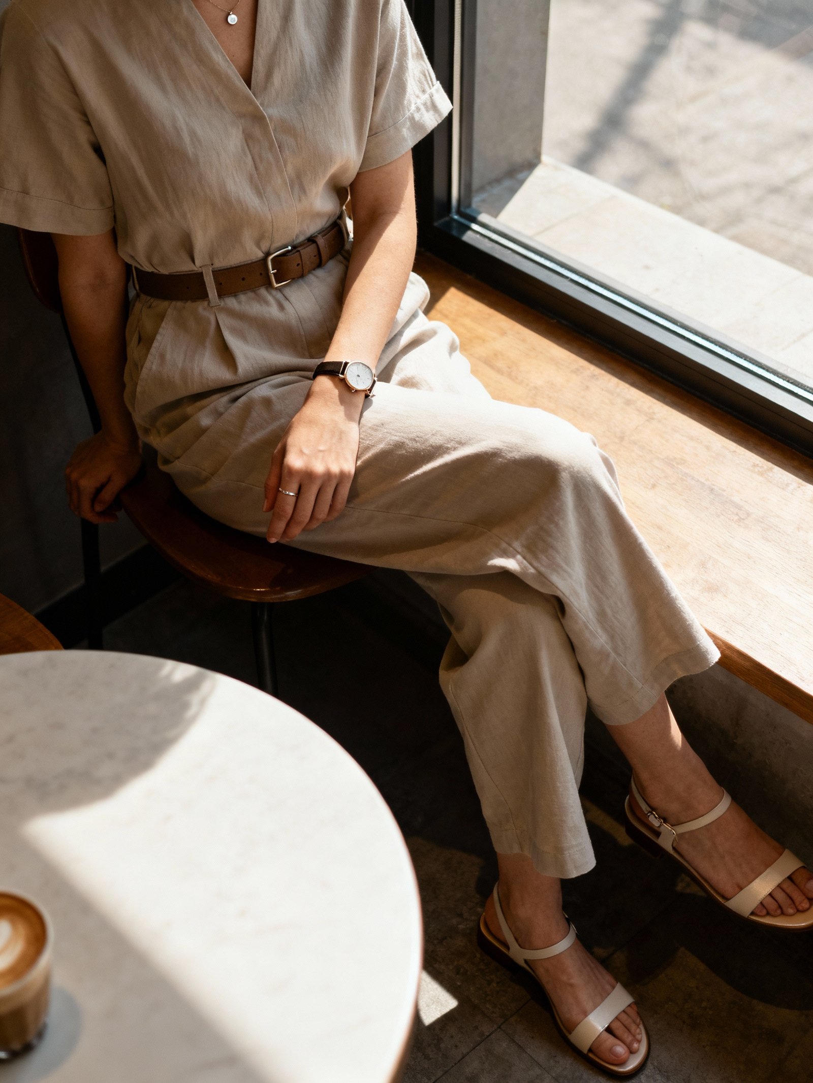 iPhone cafe-table selfie angled down of a woman wearing a minimal jumpsuit cinched with a belt, clean accessories (simple studs, slim watch) and sleek sandals, face not visible, sitting by window in coffee shop, morning natural light, natural iPhone photo quality.