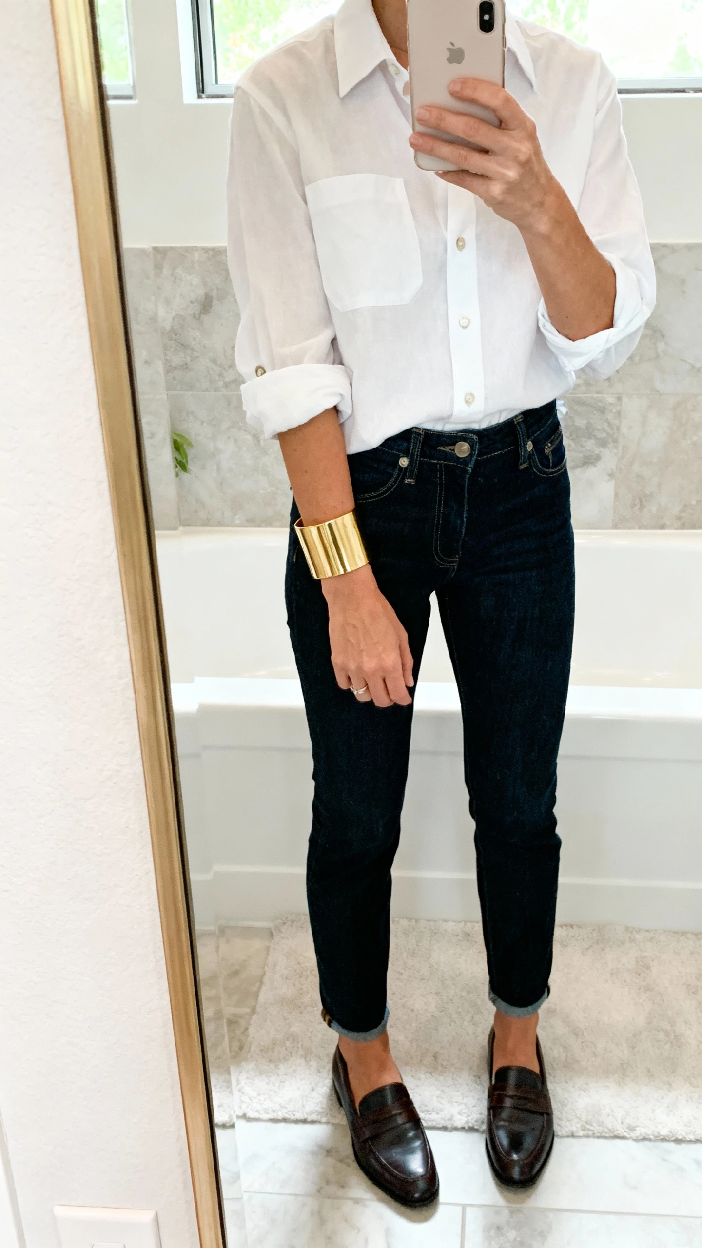 iPhone back-of-hand mirror selfie featuring a wide gold cuff bracelet peeking from a slightly pushed-up crisp white button-down sleeve, with dark denim and loafers, face not visible, bathroom mirror, bright natural light, natural iPhone photo quality