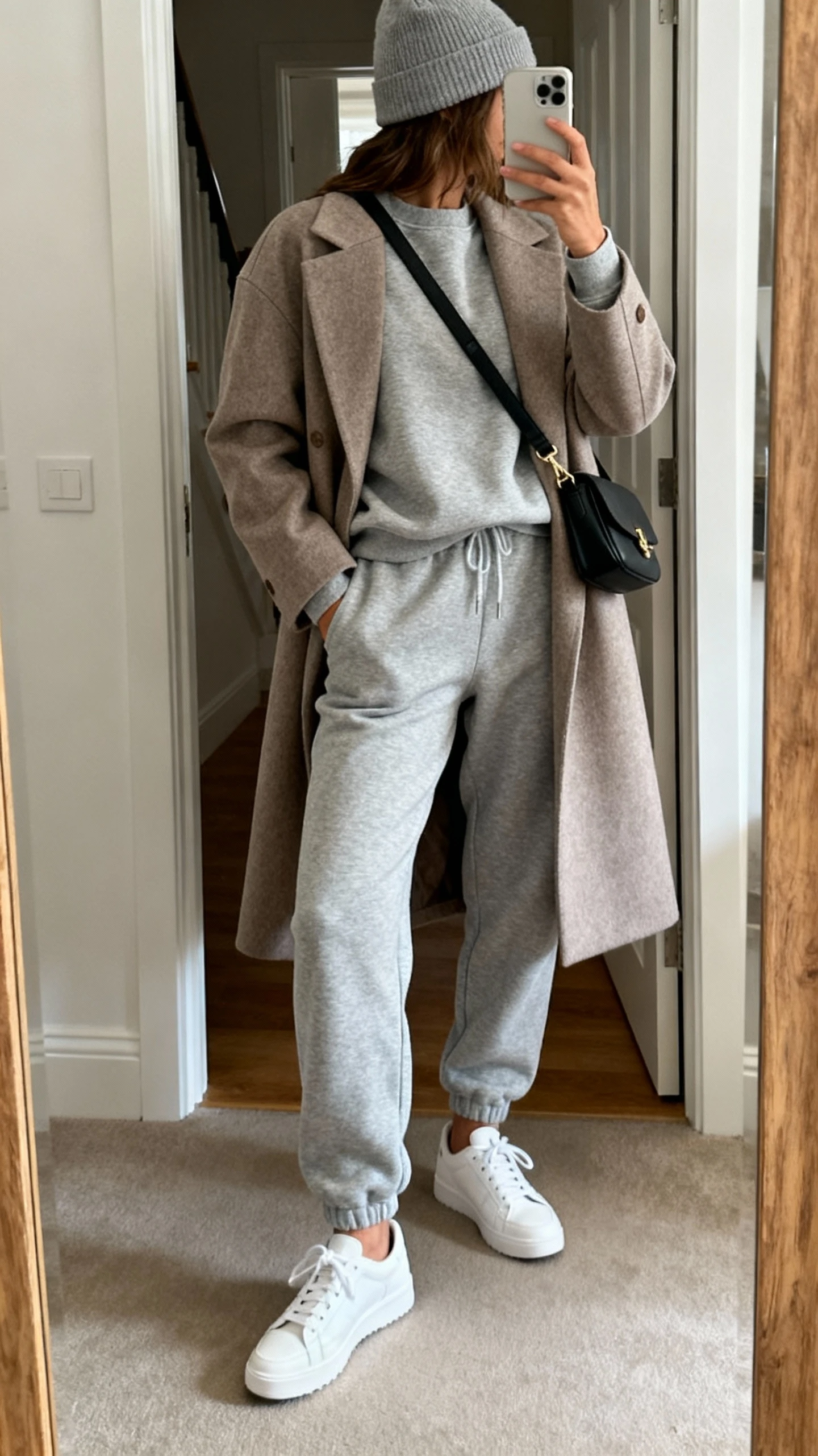 iPhone back-view mirror selfie of a woman in an elevated matching sweatsuit (crewneck and joggers) topped with a tailored topcoat, clean white sneakers, crossbody bag, and a beanie, face not visible, entryway mirror with natural daylight, casual iPhone photo.