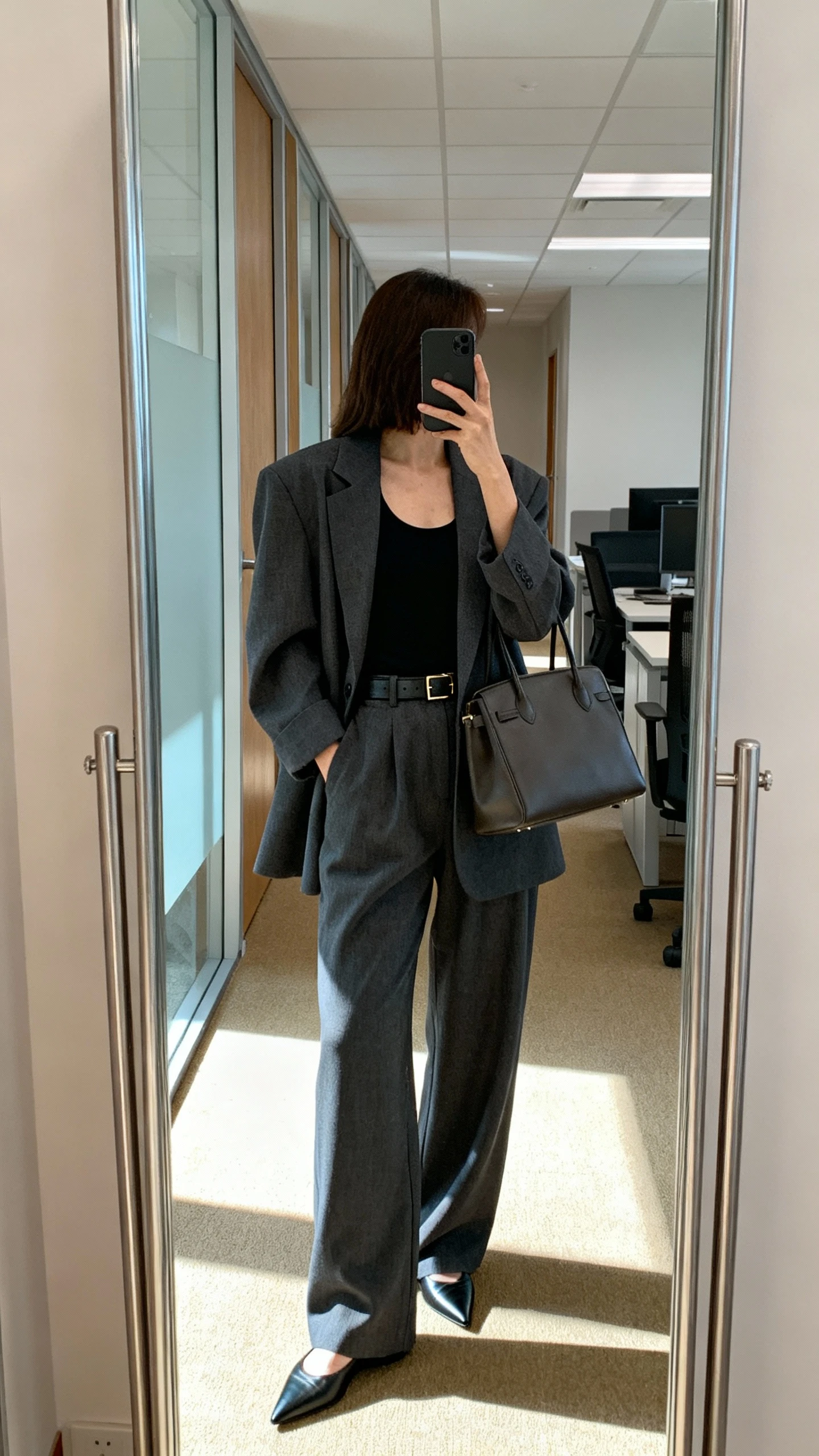 iPhone back-view mirror selfie of a woman in an oversized blazer + relaxed trouser set: slouchy charcoal blazer over matching relaxed trousers, simple black tank, pointed flats, slim leather belt, structured tote, face not visible, office hallway mirror with morning light, natural iPhone photo quality.