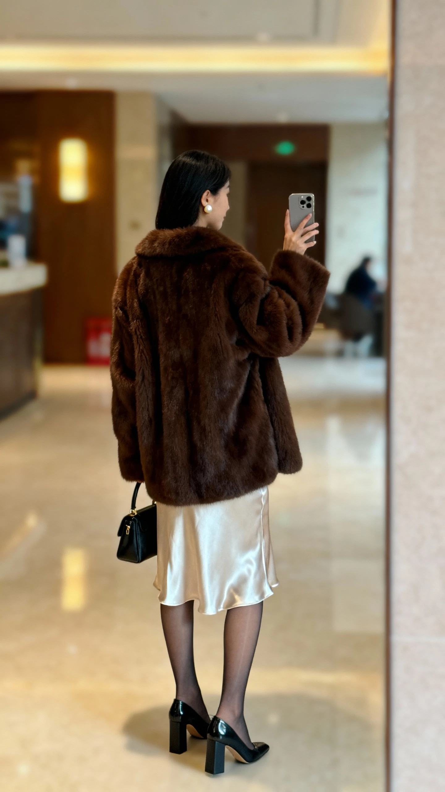 iPhone back-view selfie of a woman in a plush faux fur coat (rich chocolate brown) over a satin slip dress, sheer tights, block-heel pumps, small top-handle bag, pearl stud earrings; face not visible, lobby mirror with warm indoor lighting, casual iPhone aesthetic.