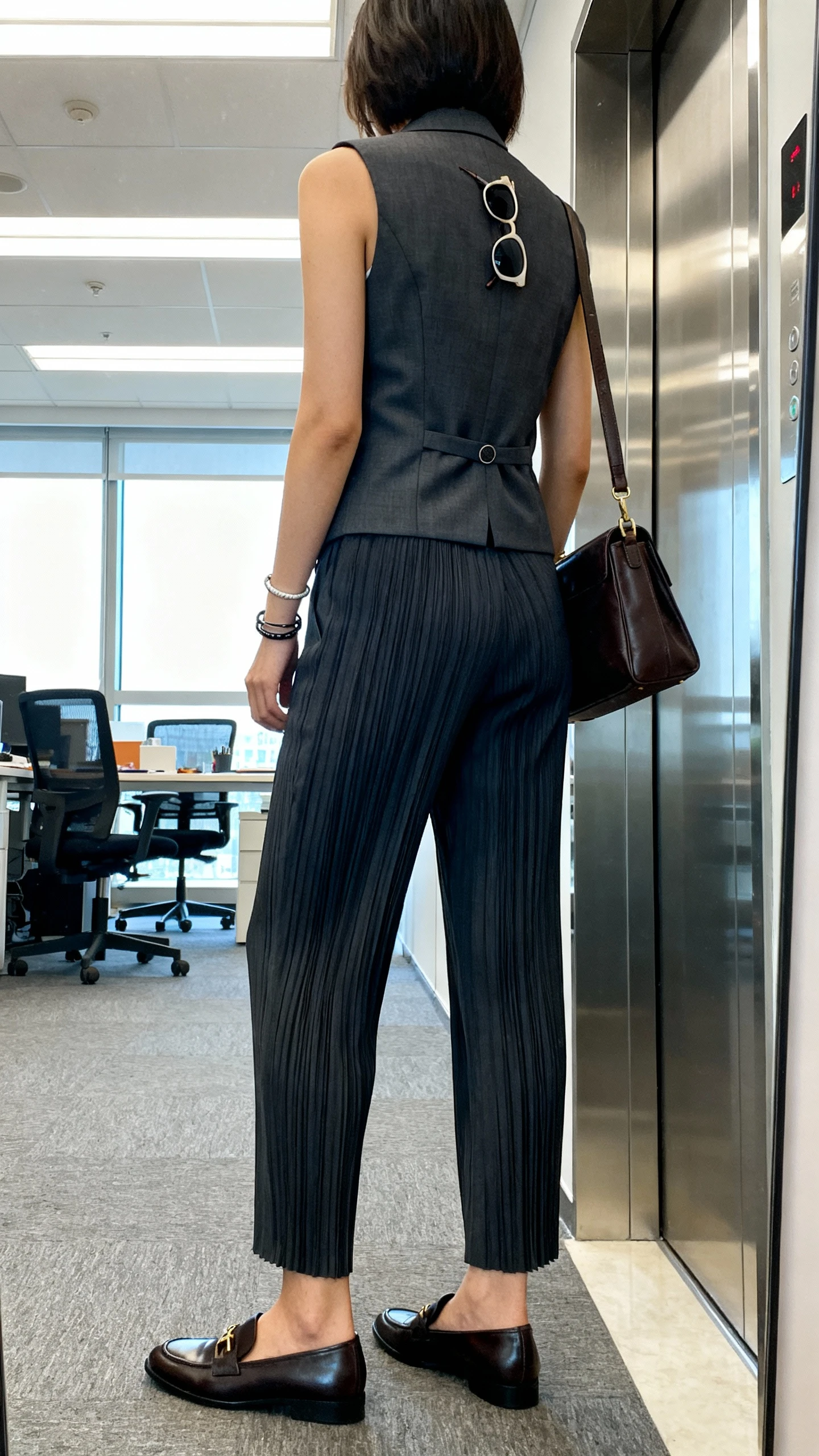 iPhone back-view selfie of a woman in a tailored vest and trousers set: fitted waistcoat with matching pleated trousers, sleek loafers, leather shoulder bag, minimalist bracelets, sunglasses hooked on vest, face not visible, elevator mirror in a creative office, bright fluorescent + window light, natural iPhone quality