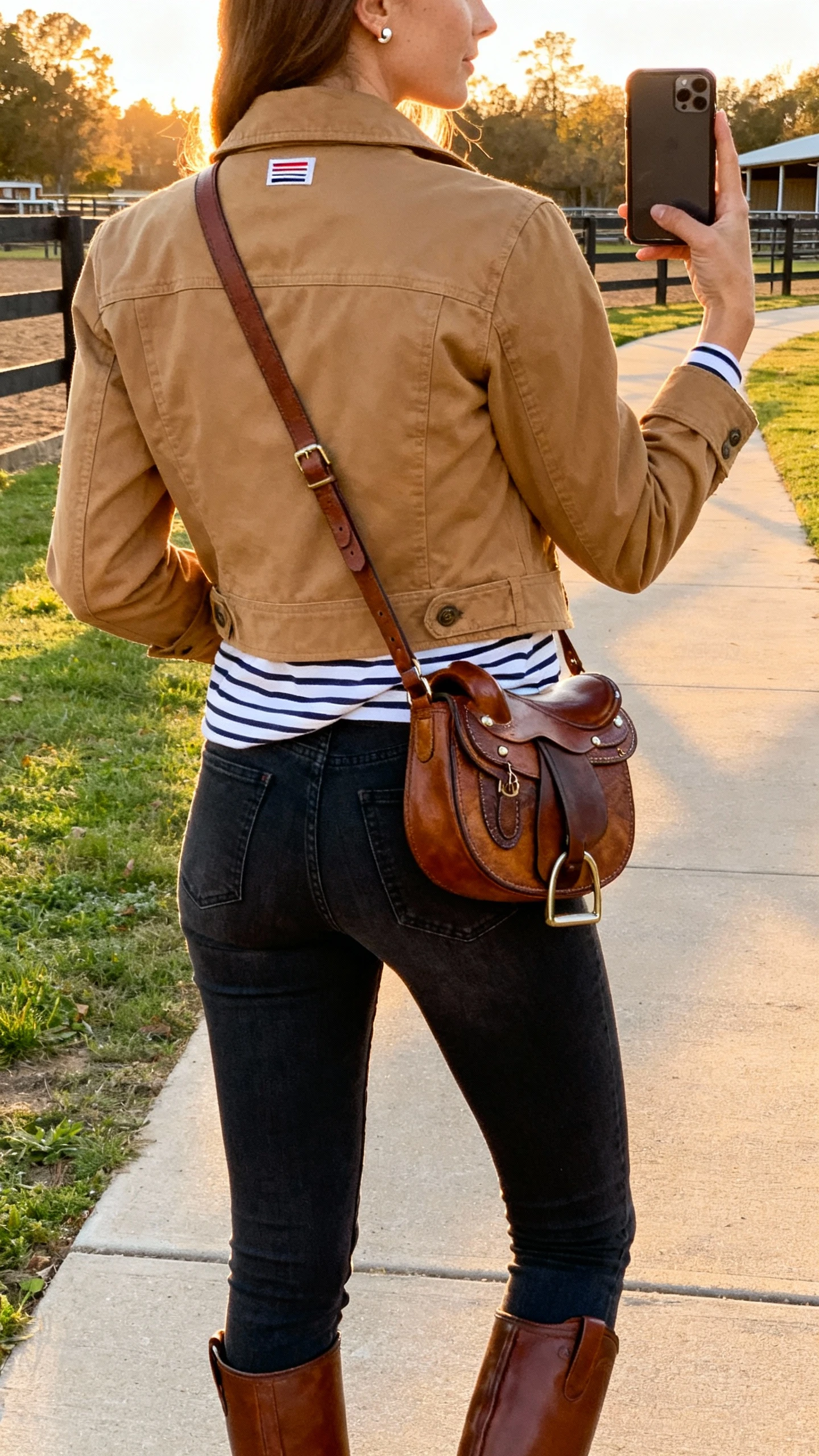 iPhone back-view selfie of a woman in an equestrian-inspired look: tailored tan field jacket, striped breton tee, high-rise dark skinny jeans, brown riding boots, leather crossbody with saddle hardware, simple stud earrings, face not visible, stable-adjacent pathway/park setting, golden late-afternoon light, casual iPhone photo.