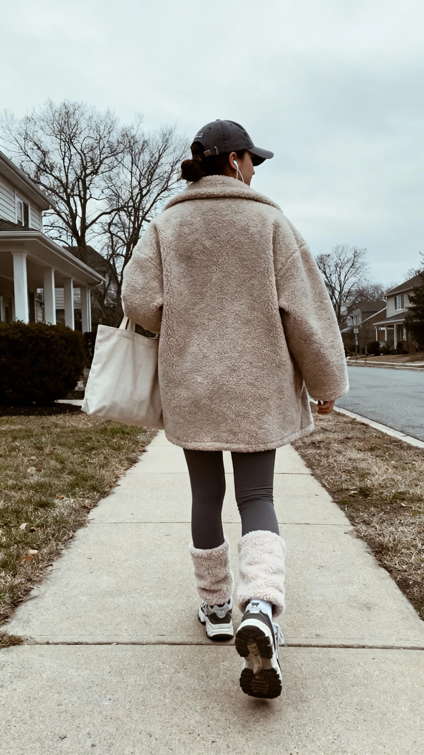iPhone back-view selfie of a woman in fleece-lined leggings, oversized wool coat, chunky sneakers, and a baseball cap with earbuds and a tote, face not visible, walking on a neighborhood sidewalk, overcast daylight, casual iPhone photo.