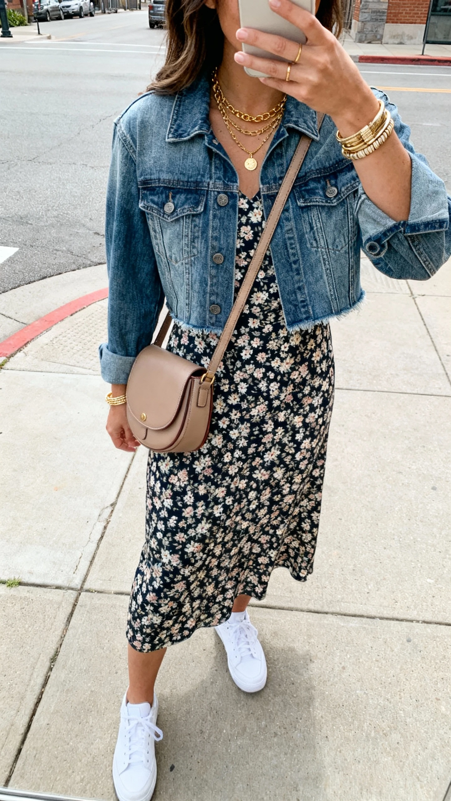 iPhone back-view selfie of a woman wearing a floral midi dress with a slightly cropped mid-wash denim jacket, white sneakers, crossbody saddle bag, layered gold necklaces and stacked bracelets; city sidewalk, natural daylight, face not visible, natural iPhone photo quality.