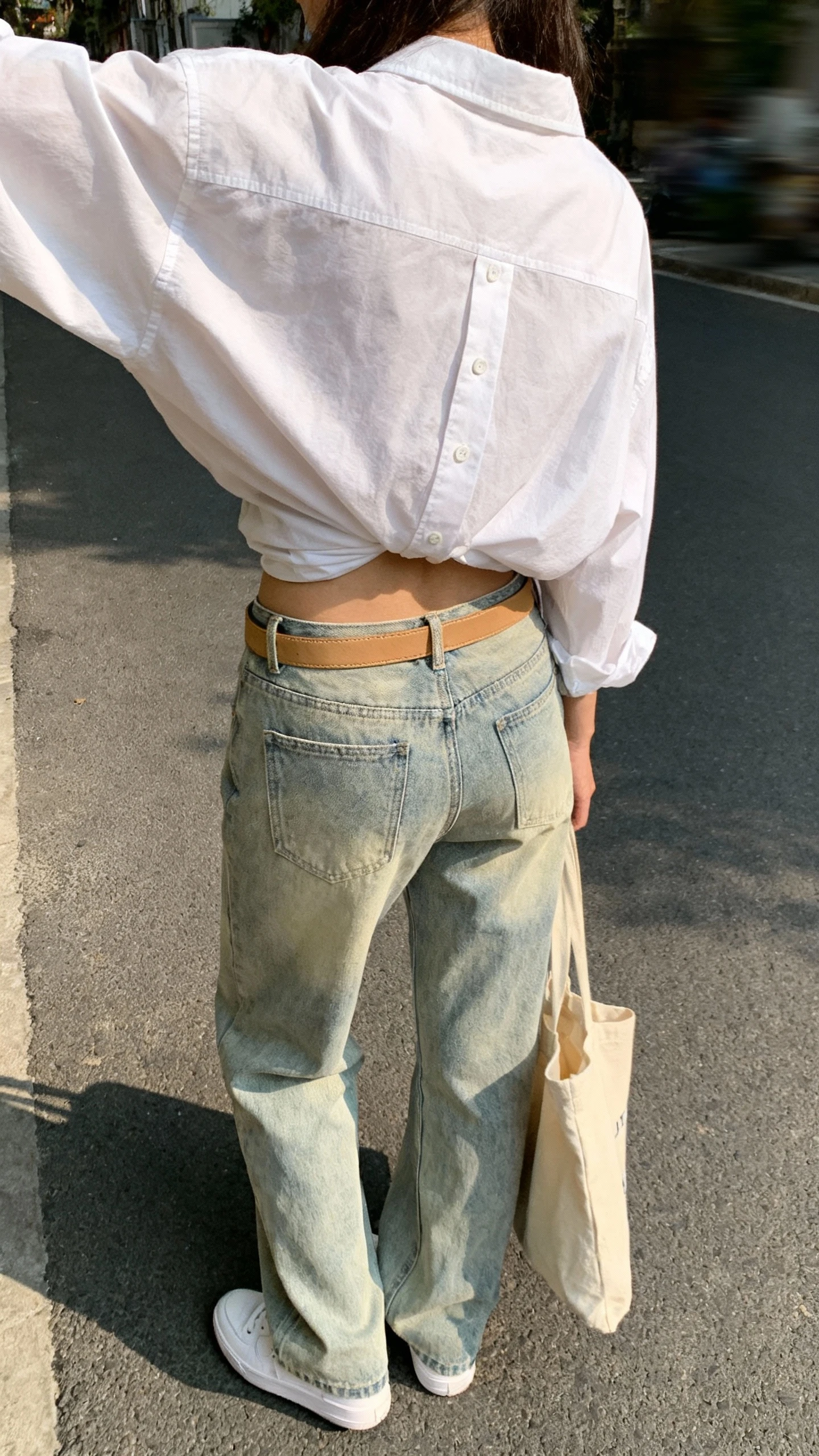 iPhone back-view selfie of a woman wearing a relaxed white button-up half-tucked into light-wash relaxed denim, tan belt, white sneakers, and a canvas tote, face not visible, taken on a quiet street with afternoon daylight, natural iPhone quality.