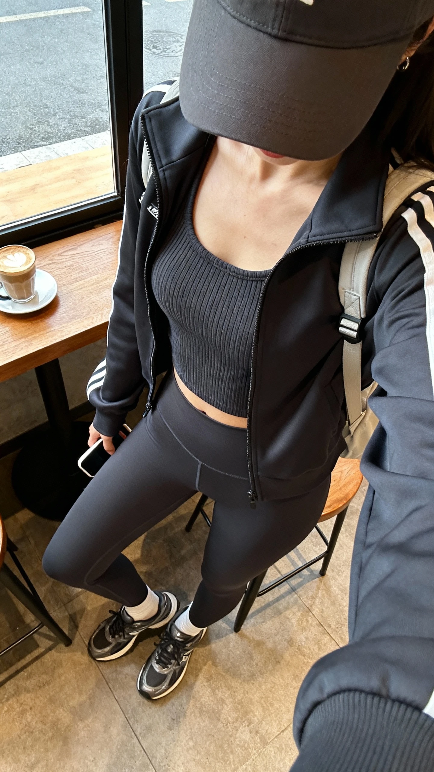 iPhone cafe table selfie (downward angle) of a woman in elevated athleisure: tailored track jacket, ribbed tank, high-rise leggings, sleek trainers, baseball cap, and a minimal backpack, face not visible, cozy coffee shop window light, natural iPhone photo quality.