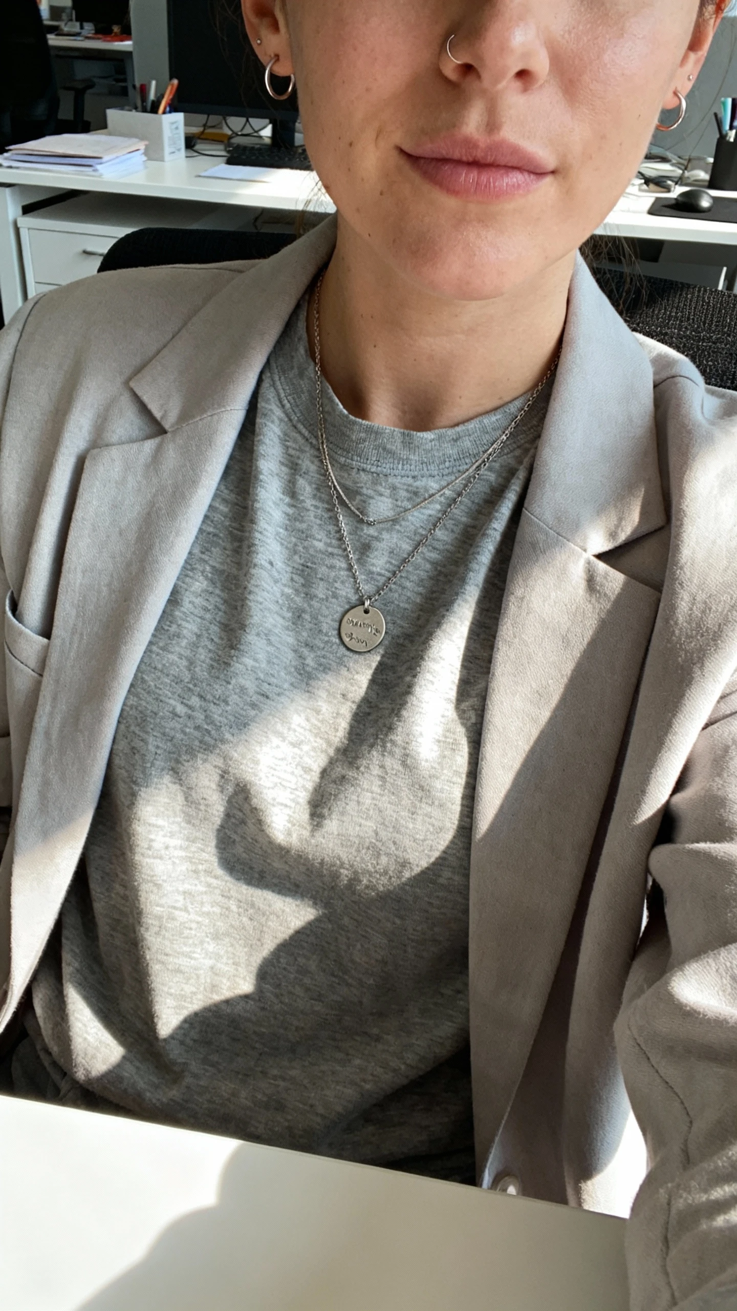 iPhone chest-up selfie of a woman wearing an everyday personal pendant necklace (small engraved disc) layered over a heather gray crewneck tee and lightweight blazer, tiny hoops, face not visible, office desk setting with morning light, natural iPhone photo quality