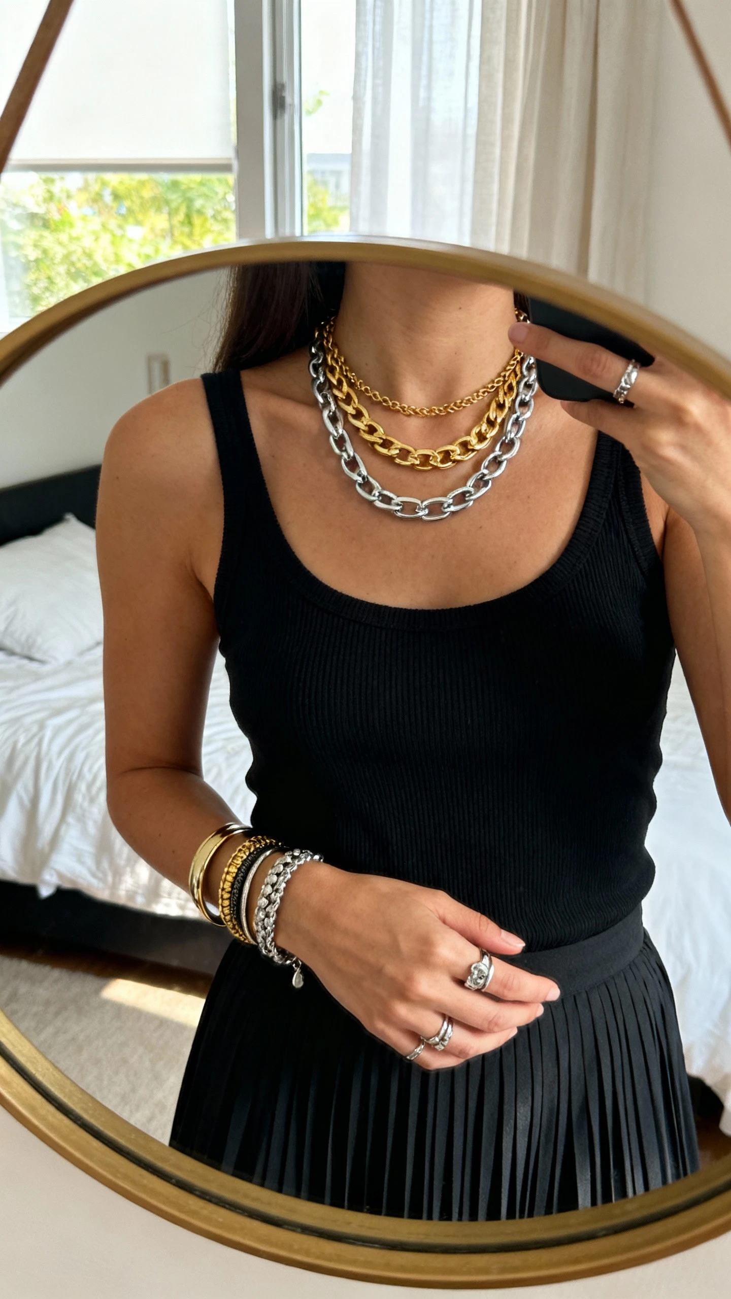 iPhone close-up mirror selfie of a woman wearing a mixed-metal stack: gold and silver layered necklaces plus mixed bangles and rings, with a black ribbed tank and pleated skirt, face not visible, bedroom mirror, morning window light, natural iPhone photo quality