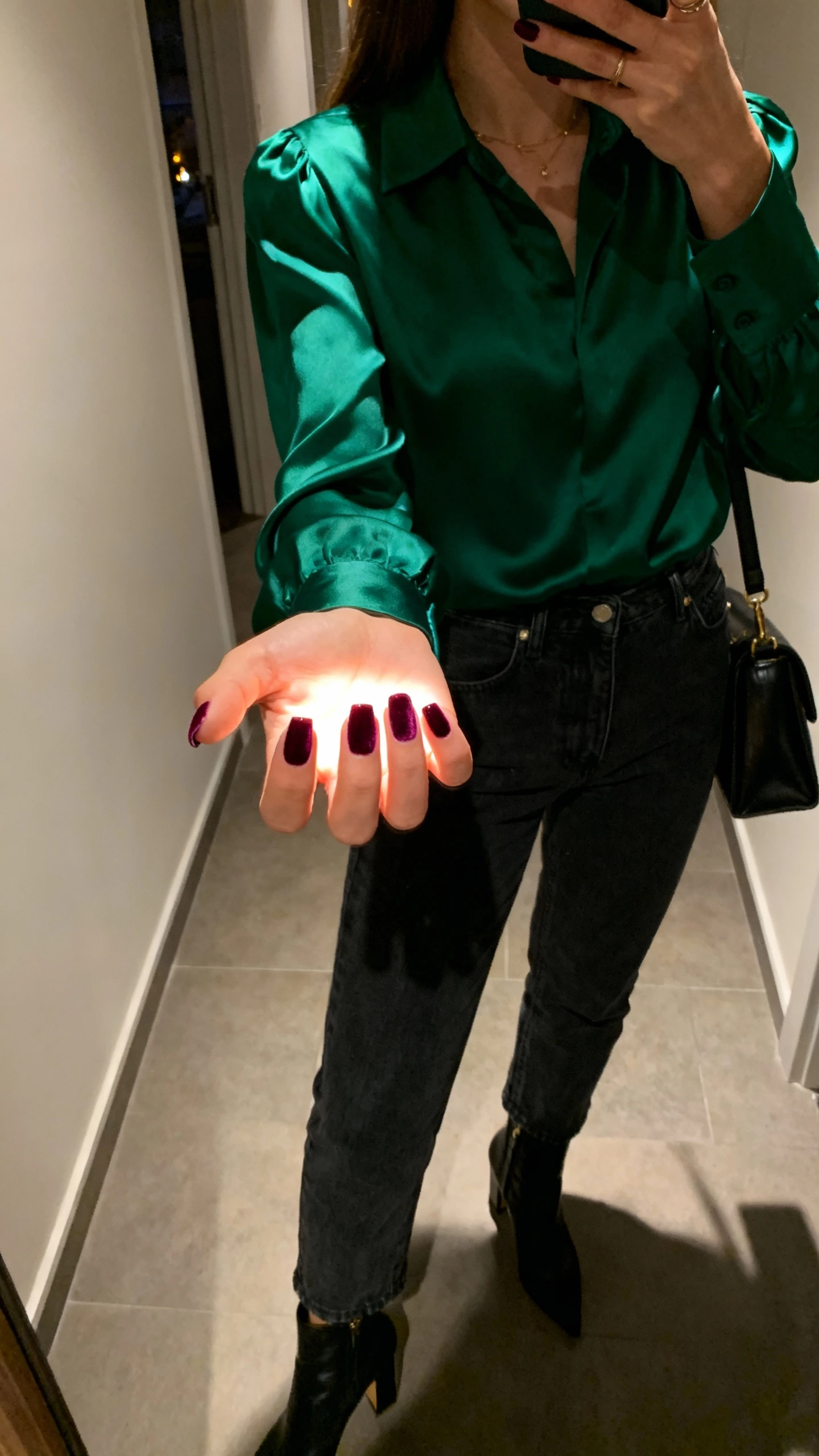 iPhone close-up selfie of a woman’s hand catching the light to show velvet sheen cat-eye nails, wearing a satin blouse in deep green, dark denim, heeled ankle boots, and a structured shoulder bag; evening indoor hallway light, face not visible, casual iPhone shot.
