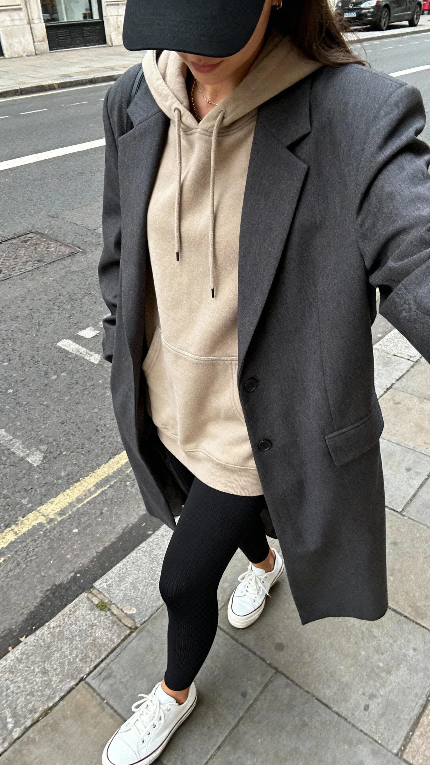 iPhone cropped-at-shoulders selfie of a woman wearing black thick sculpting leggings, sand longline hoodie, charcoal oversized blazer, clean white leather sneakers, black baseball cap; face not visible, walking on a city street, natural daylight, natural iPhone photo quality.