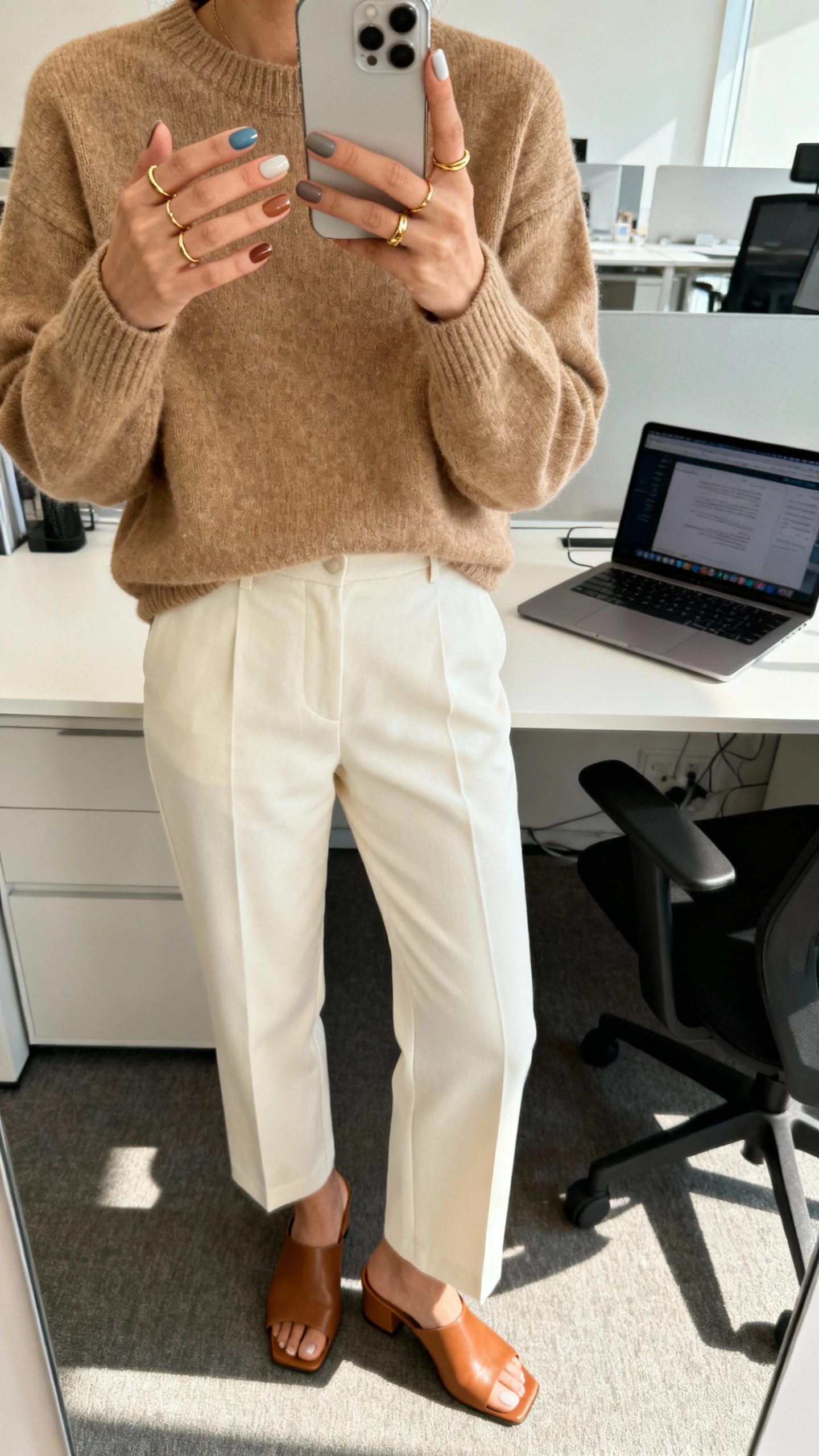 iPhone desk selfie cropped at shoulders showing soft skittle shades in all neutral tones on nails, wearing a camel knit, tailored cream trousers, dainty gold rings, and tan mules; office setting with laptop, morning light, face not visible, natural iPhone aesthetic.