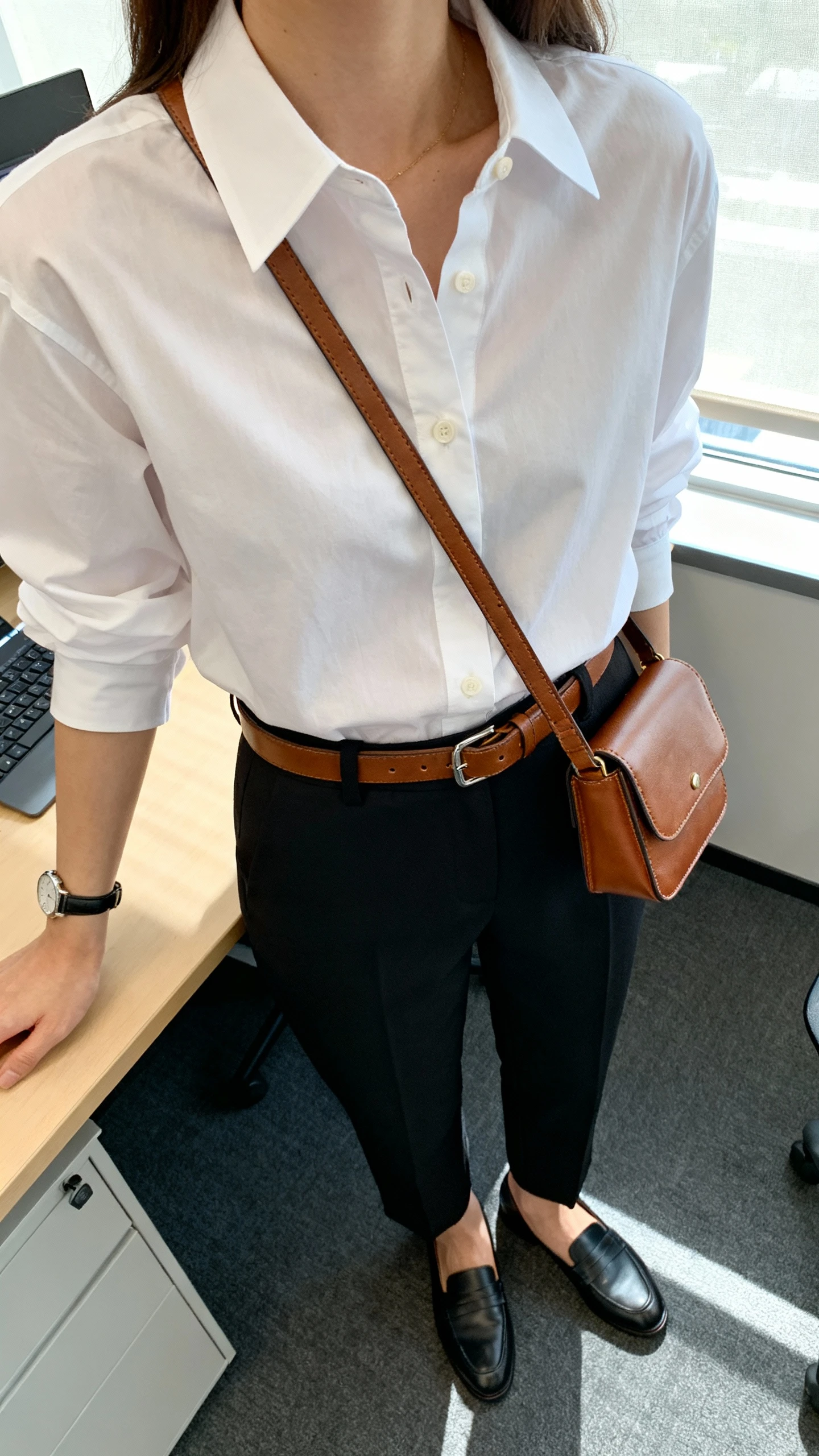 iPhone desk-side selfie cropped at neck of a woman wearing a crisp white button-down with leather-feel belt and leather-feel mini crossbody, black tailored trousers, loafers, slim watch, face not visible, office setting with window light, natural iPhone quality.