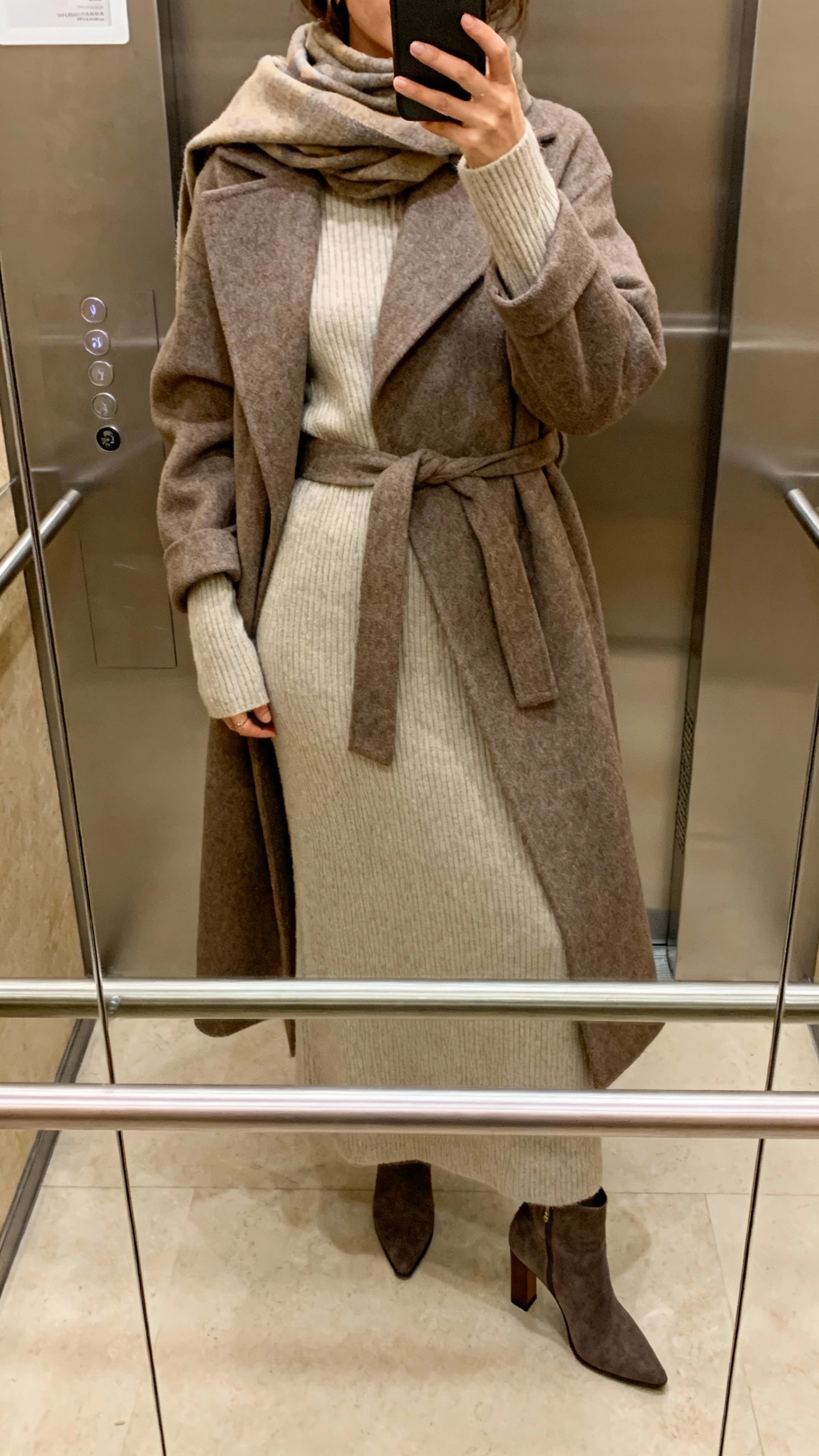 iPhone elevator mirror selfie of a woman in a cozy long-sleeve knit maxi dress with a belted wool coat, heeled ankle boots, scarf, face not visible, warm indoor lighting, natural iPhone photo quality.
