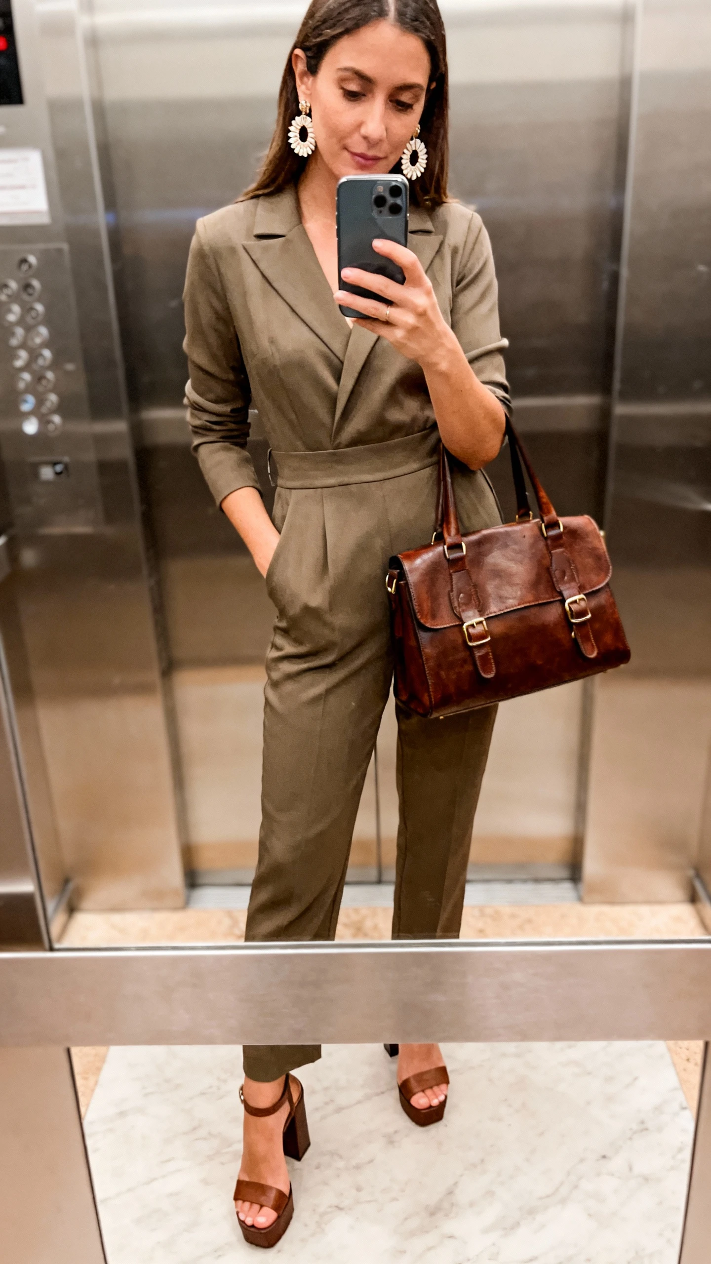 iPhone elevator mirror selfie of a woman in a tailored power jumpsuit with a defined waist, block-heel sandals, statement earrings, leather satchel, face not visible, bright indoor lighting, candid iPhone shot.