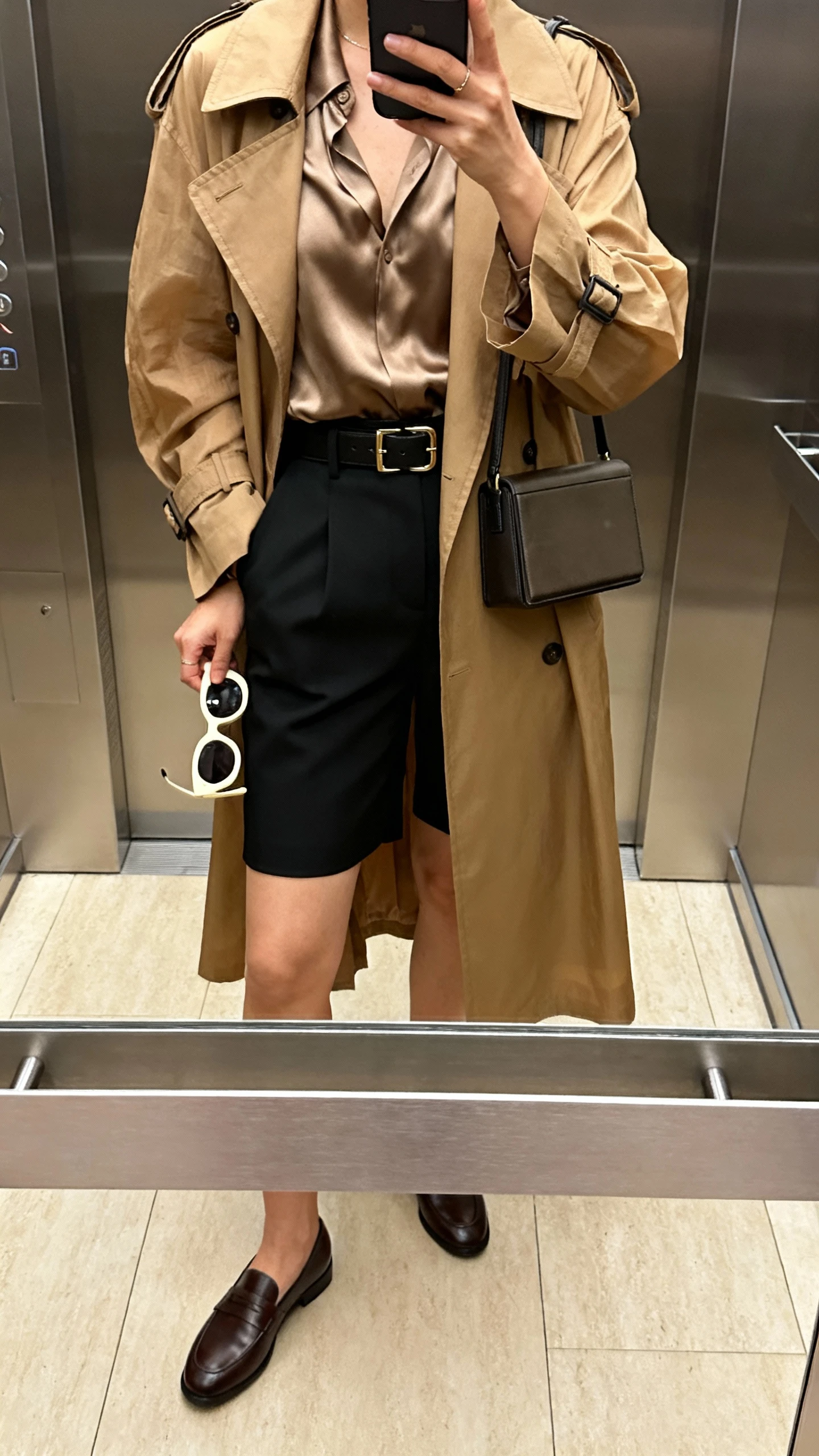 iPhone elevator mirror selfie of a woman in a tan lightweight trench over tailored black shorts, tucked silky blouse, loafers, structured belt, boxy crossbody, statement sunglasses in hand; indoor lighting, face not visible, cropped at shoulders, natural iPhone photo quality.