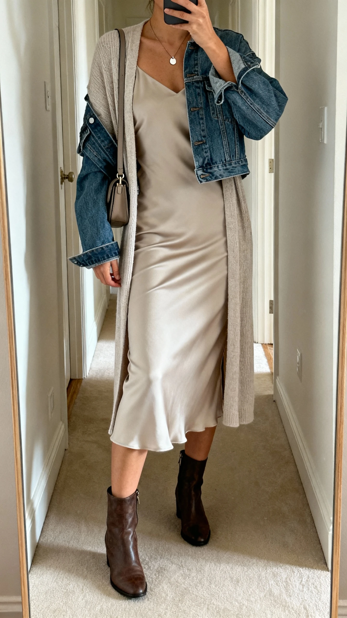 iPhone hallway mirror selfie of a woman in a bias-cut slip dress layered with a lightweight cardigan and a cropped denim jacket, ankle boots, simple pendant, and a small shoulder bag, face not visible, natural morning light, casual iPhone aesthetic.