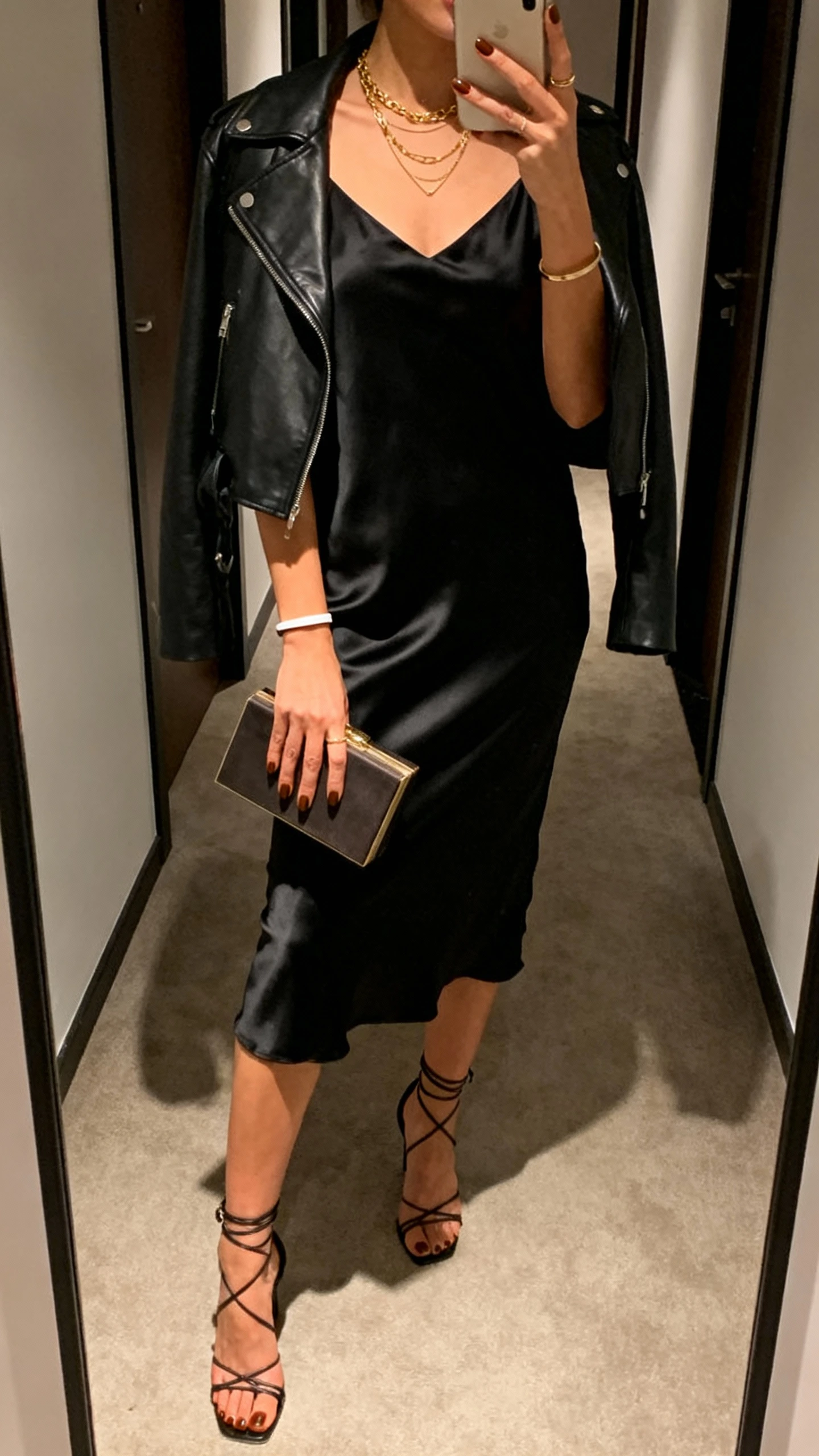 iPhone hallway mirror selfie of a woman in a black satin slip dress with a cropped leather jacket, strappy heeled sandals, espresso-brown manicure, delicate layered gold necklaces and a slim cuff bracelet, small box clutch; face not visible, cropped at neck, evening indoor lighting, natural iPhone quality.