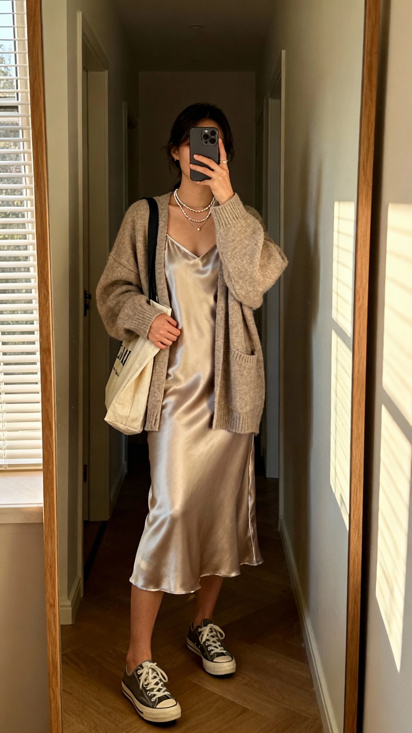 iPhone hallway mirror selfie of a woman in an oversized cardigan layered over a satin slip dress with classic sneakers, delicate layered necklaces, tote bag, face not visible, full-length mirror, afternoon window light, natural iPhone photo quality.