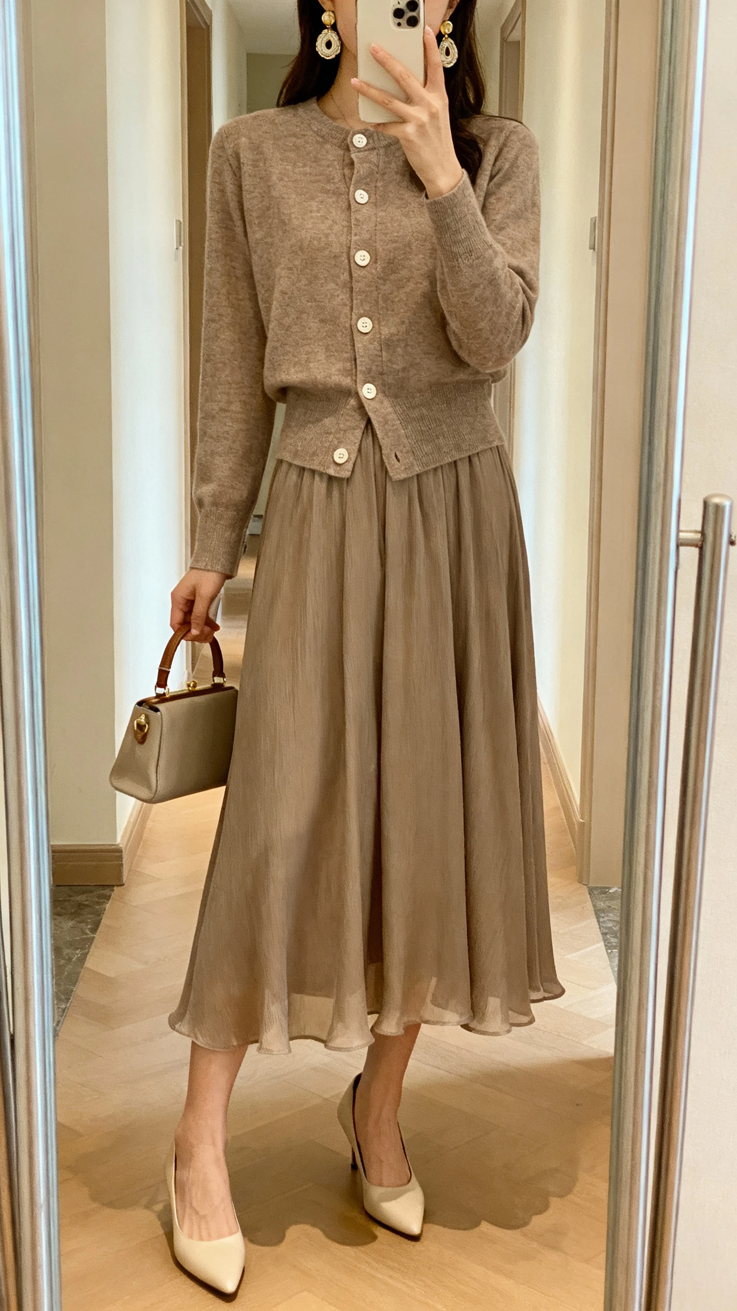 iPhone hallway mirror selfie of a woman wearing a flowy midi skirt with a matching cardigan set (cardigan buttoned as a top), kitten heels, vintage-inspired earrings, small top-handle bag, face not visible, warm indoor lighting, casual iPhone photo.
