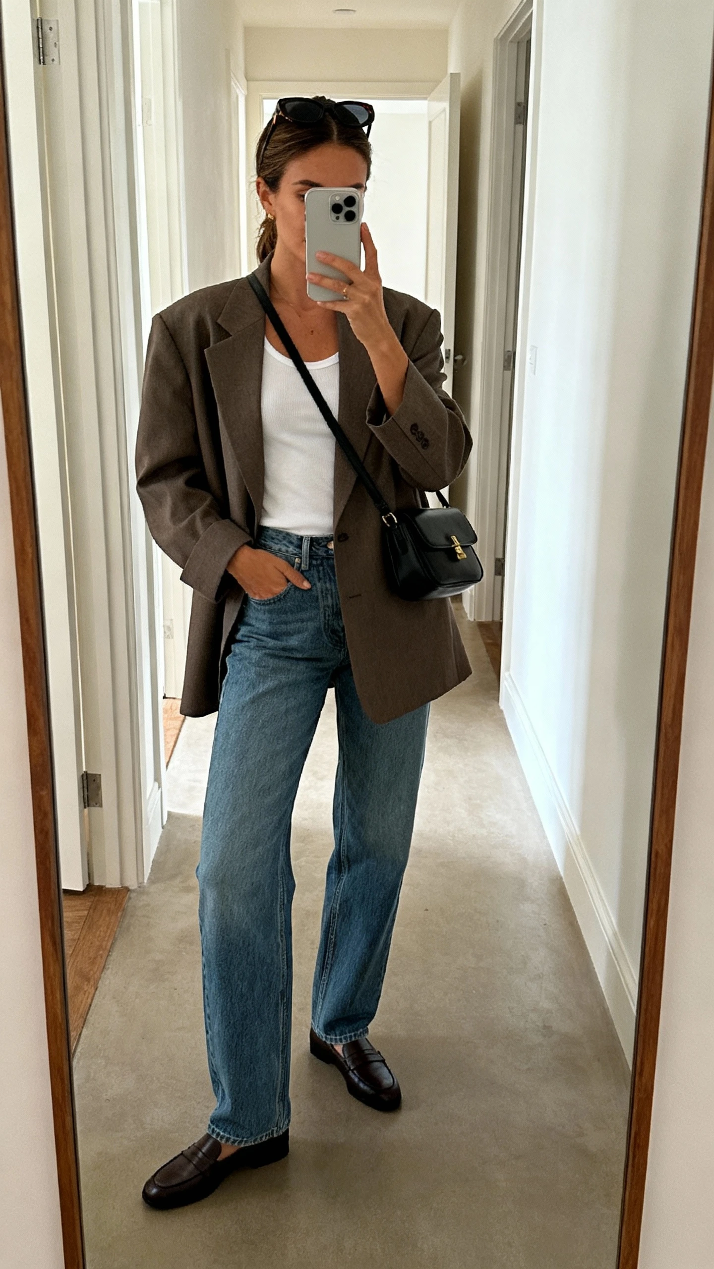 iPhone hallway mirror selfie of a woman wearing an oversized blazer over straight-leg blue jeans, classic white tank, loafers, black crossbody bag, sunglasses pushed into hair, face not visible, indoor lighting, casual iPhone aesthetic.