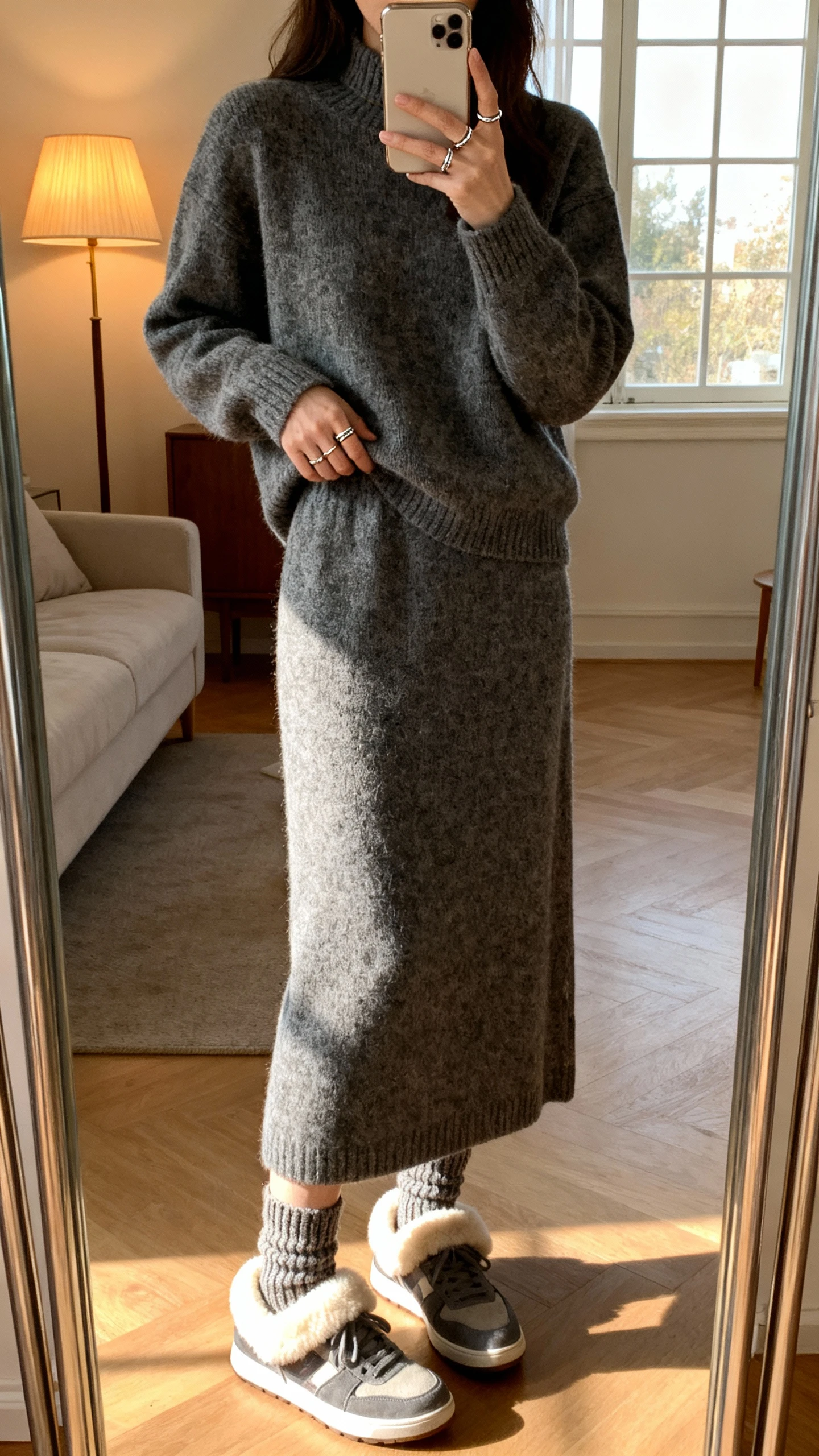 iPhone living-room mirror selfie of a woman in a matching sweater skirt set with shearling-lined sneakers, knit socks peeking, delicate ring stack, face not visible, warm indoor lamp + window light, natural iPhone quality.