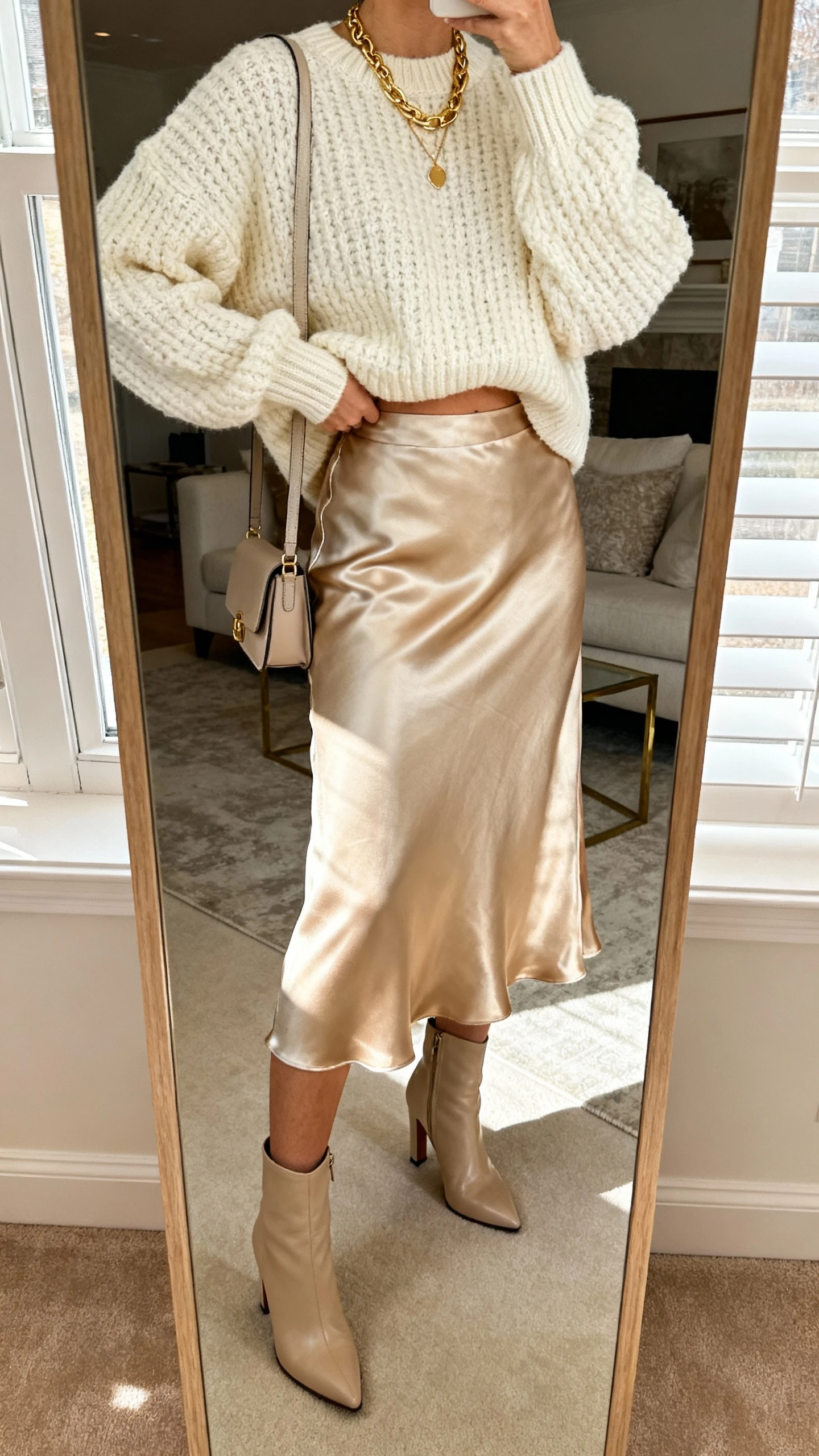 iPhone mirror selfie cropped at shoulders of a woman in a cozy sweater + midi skirt: chunky cream sweater half-tucked into a satin midi skirt in champagne, heeled ankle boots, layered gold necklaces, small shoulder bag, face not visible, living room mirror with soft window light, natural iPhone photo quality.