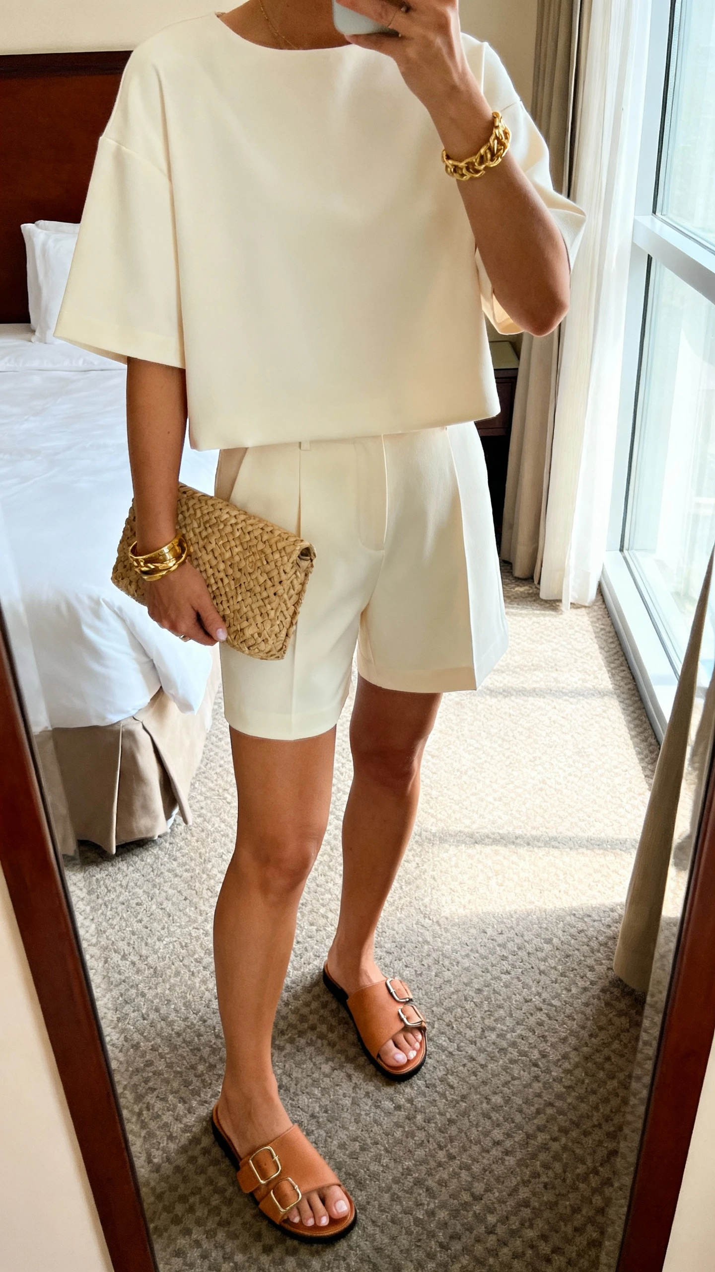 iPhone mirror selfie of a woman in a chic matching set: cream short-sleeve boxy top with matching tailored shorts, tan slide sandals, woven clutch, gold bracelets; hotel room mirror, face not visible, natural window light, casual iPhone quality.