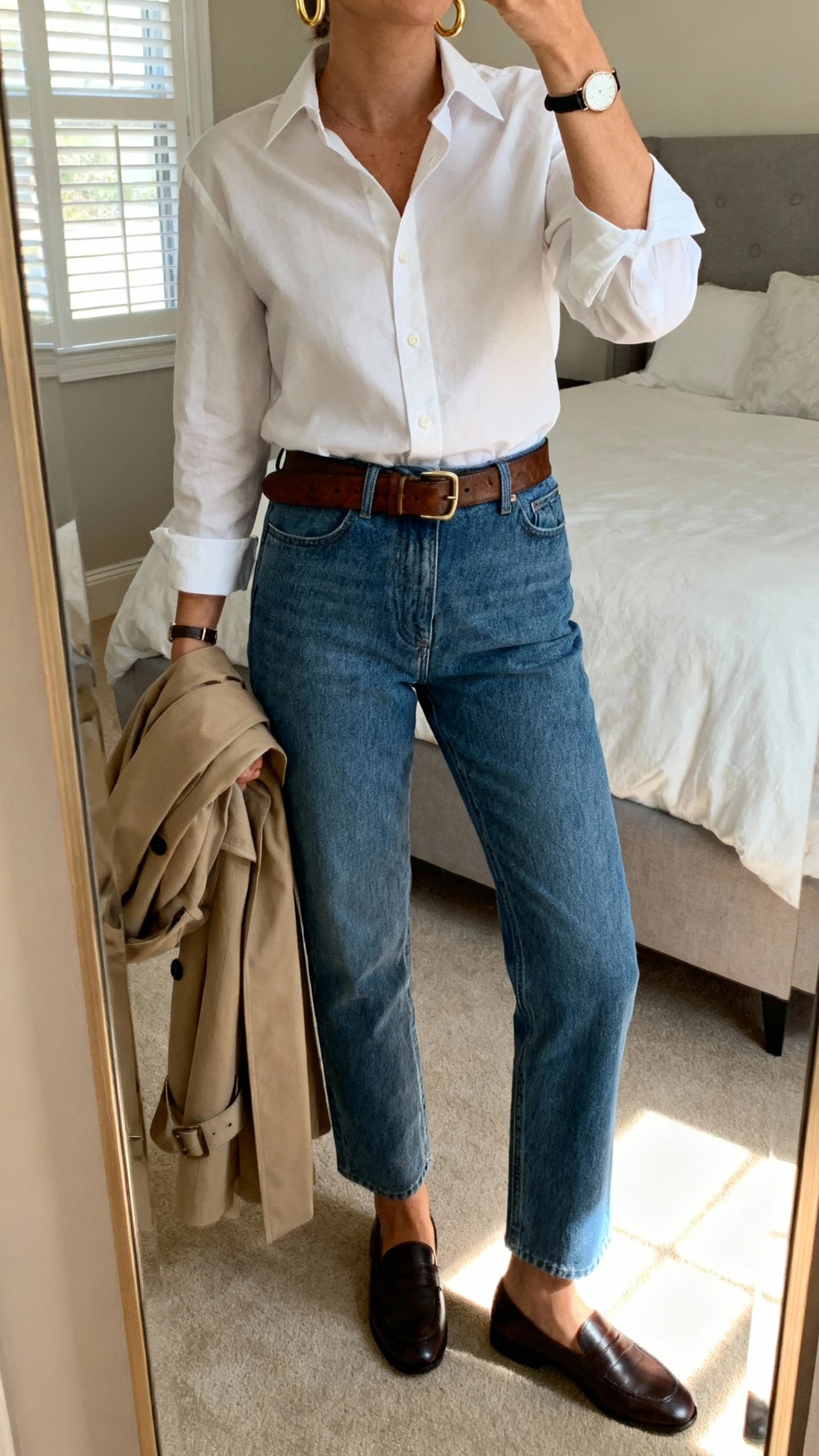 iPhone mirror selfie of a woman in a crisp white button-down tucked into straight-leg blue jeans, leather belt, loafers, gold hoop earrings, simple watch, optional trench draped on arm; face not visible, cropped at neck, bedroom mirror with morning window light, natural iPhone photo quality.