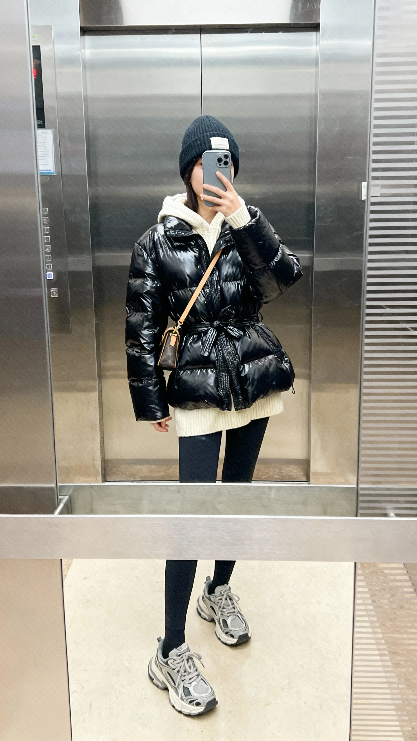iPhone mirror selfie of a woman in a glossy black puffer with cinched waist, cream knit hoodie peeking out, leggings, chunky dad sneakers, crossbody bag, beanie; face not visible, elevator mirror, bright indoor lighting, casual iPhone aesthetic.