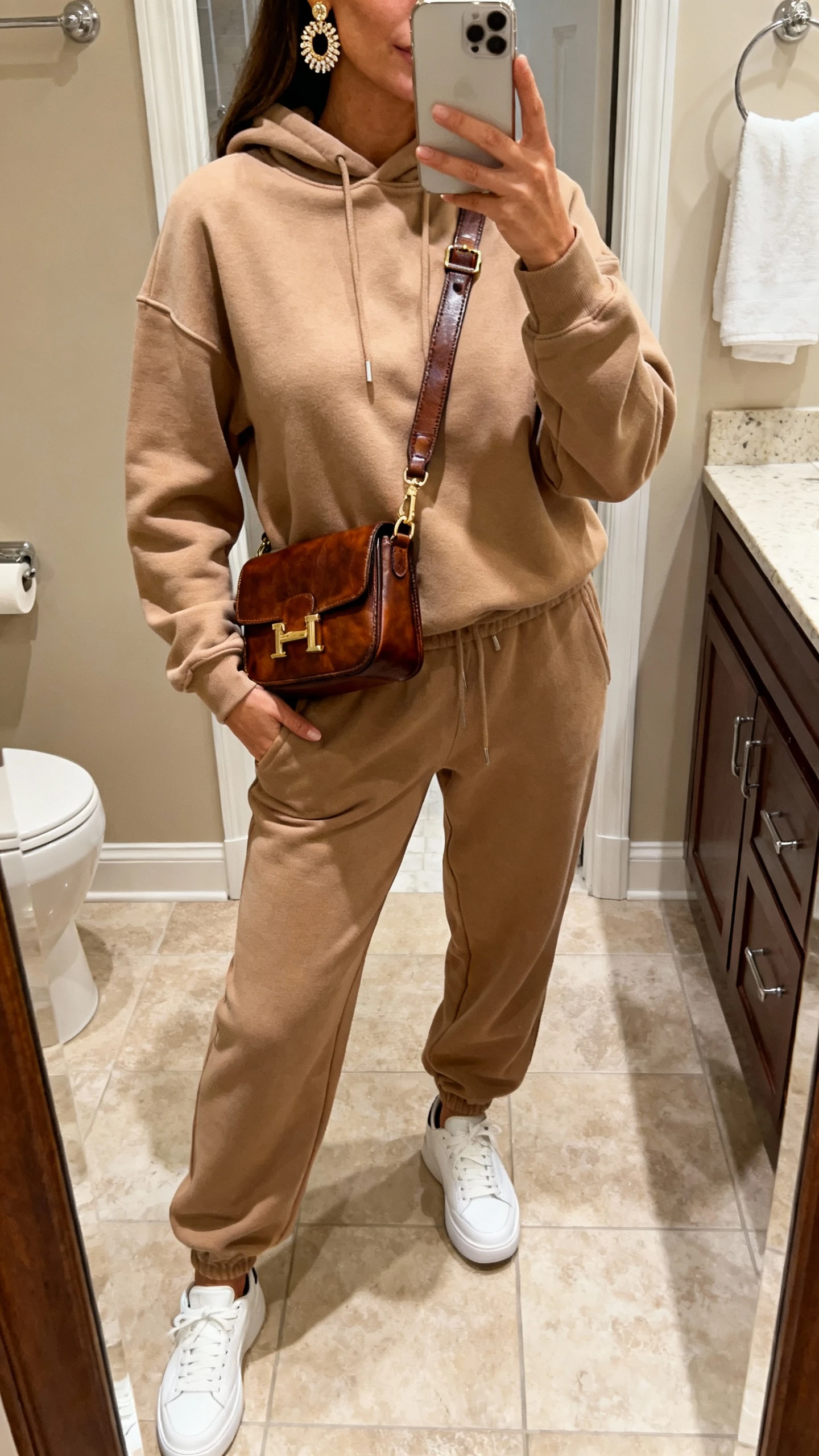 iPhone mirror selfie of a woman in a luxe sweatsuit: matching premium joggers and hoodie, styled with a structured leather crossbody, statement earrings, and polished white sneakers, face not visible, bathroom mirror, warm indoor lighting, natural iPhone photo.
