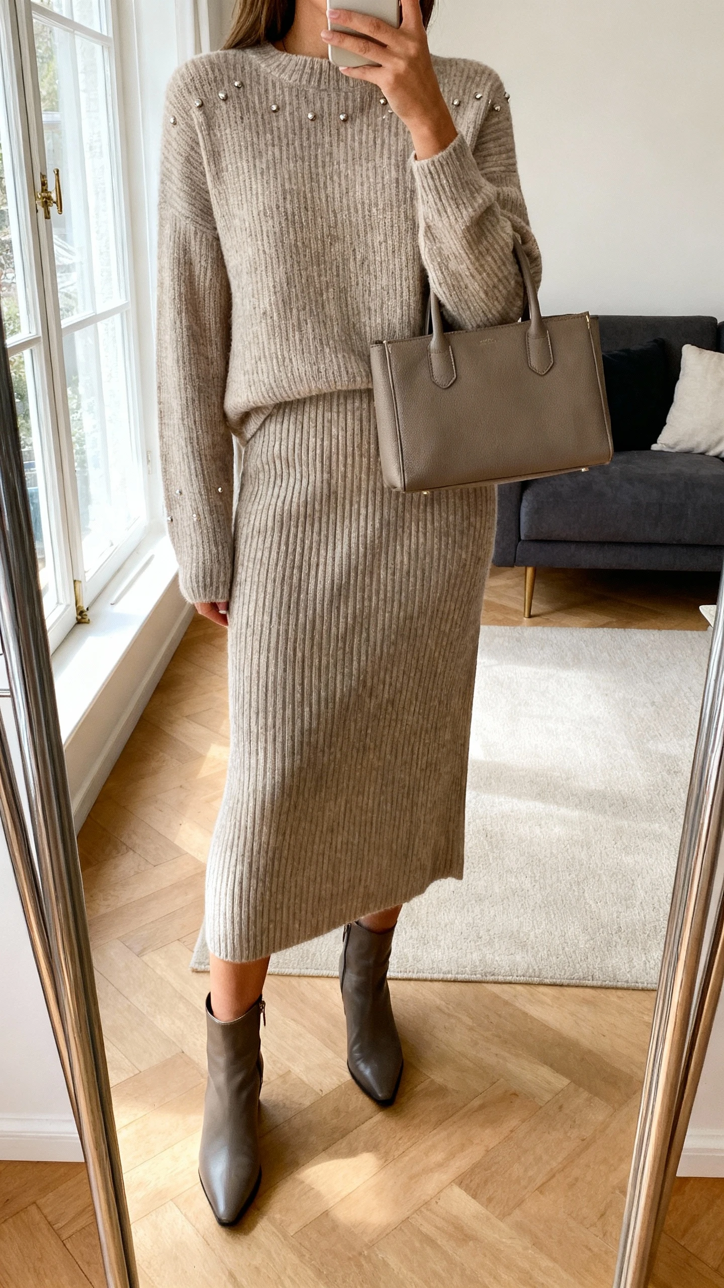 iPhone mirror selfie of a woman in a matching knit set (crewneck sweater and ribbed midi skirt), sleek ankle boots, minimal studs, structured top-handle bag, face not visible, cropped at shoulders, living room mirror with soft window light, natural iPhone quality.