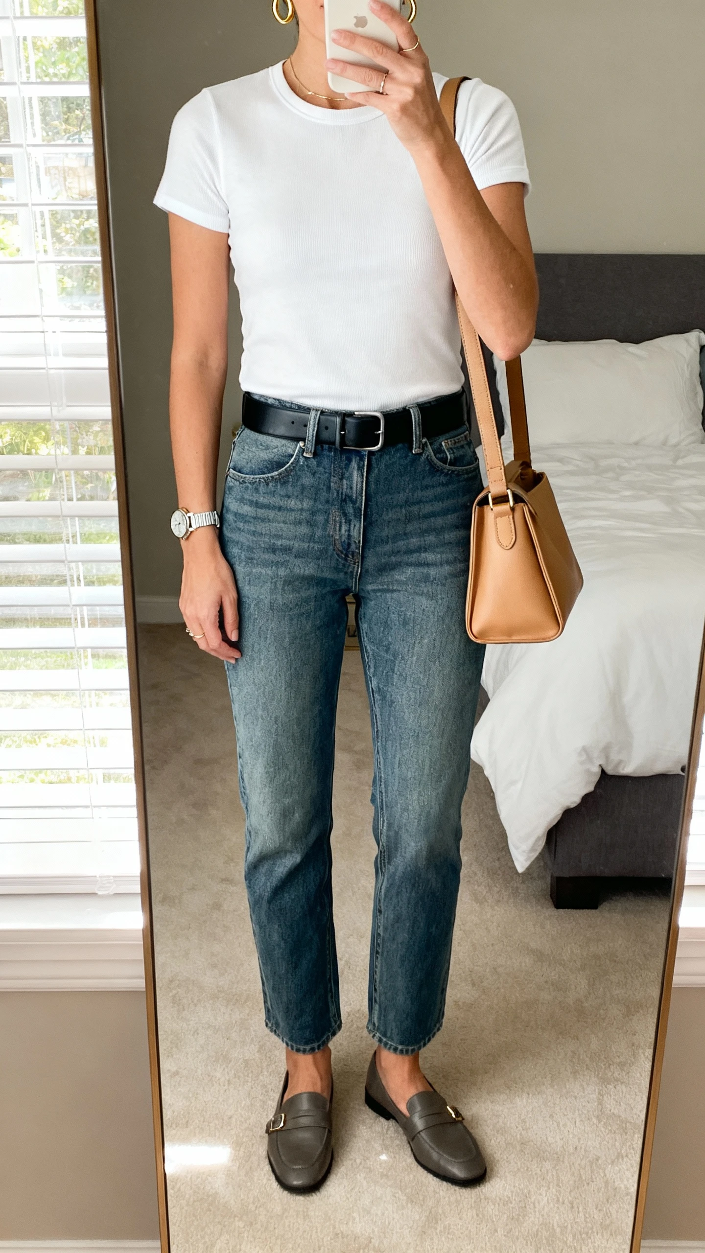 iPhone mirror selfie of a woman in a polished tee-and-jeans glow-up: fitted white crew tee tucked into straight-leg mid-wash jeans, sleek black belt, loafers, delicate gold hoops and watch, structured shoulder bag in camel, face not visible, cropped at neck, bedroom mirror with morning window light, natural iPhone photo quality.