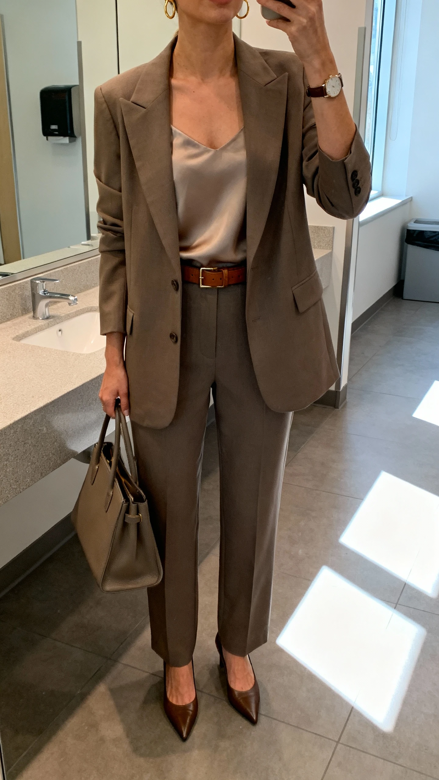 iPhone mirror selfie of a woman in a quiet-luxury blazer set: tailored blazer with matching straight-leg trousers, silk camisole, pointed-toe pumps, slim leather belt, structured tote, minimal gold hoops and watch, face not visible, cropped at neck, corporate office restroom mirror, morning window light, natural iPhone photo quality