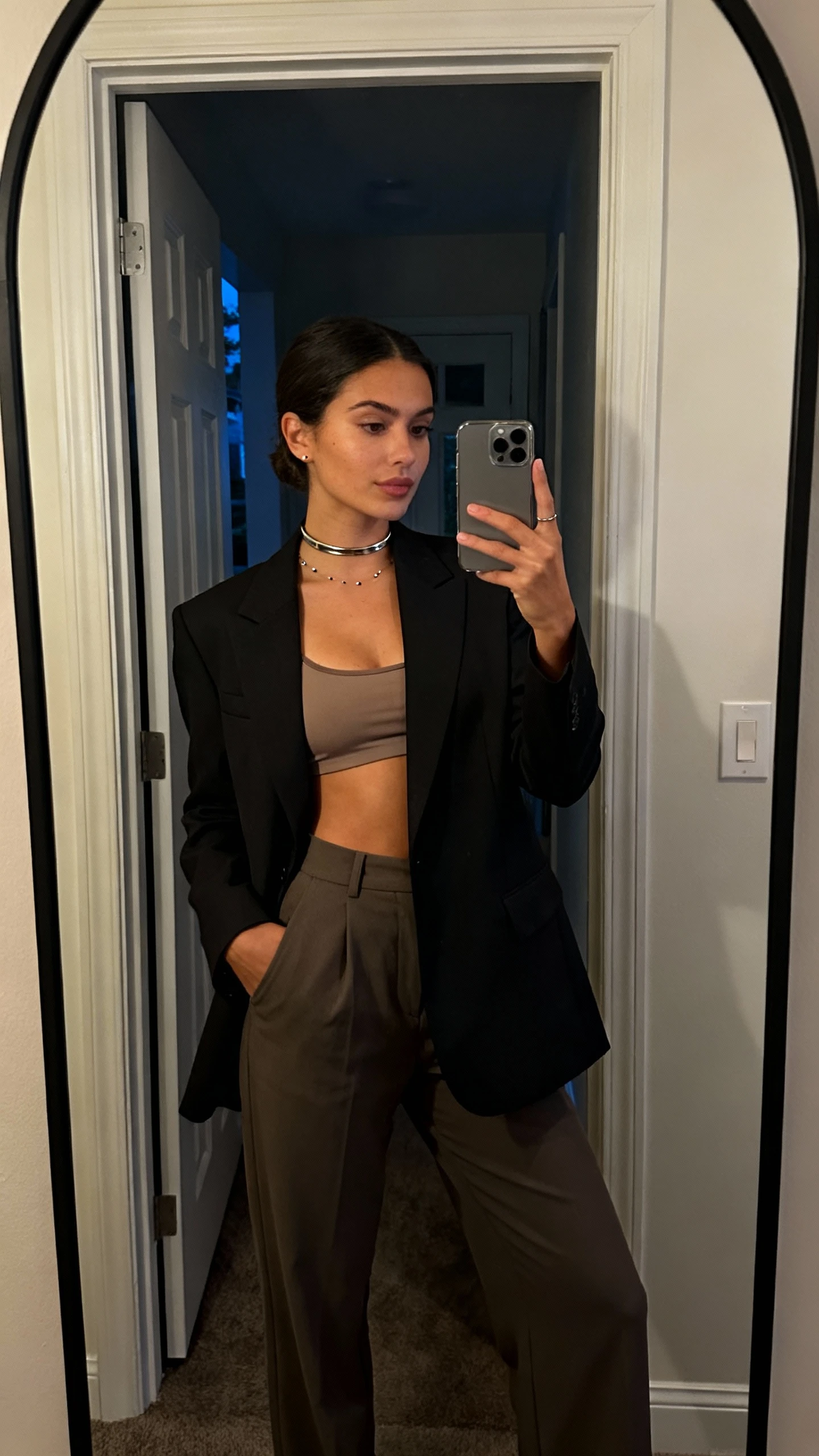 iPhone mirror selfie of a woman in a sharp blazer over a camisole pairing a slim metallic choker with tiny studs, high-waisted trousers, face not visible, full-length mirror in entryway, soft indoor evening light, natural iPhone photo quality