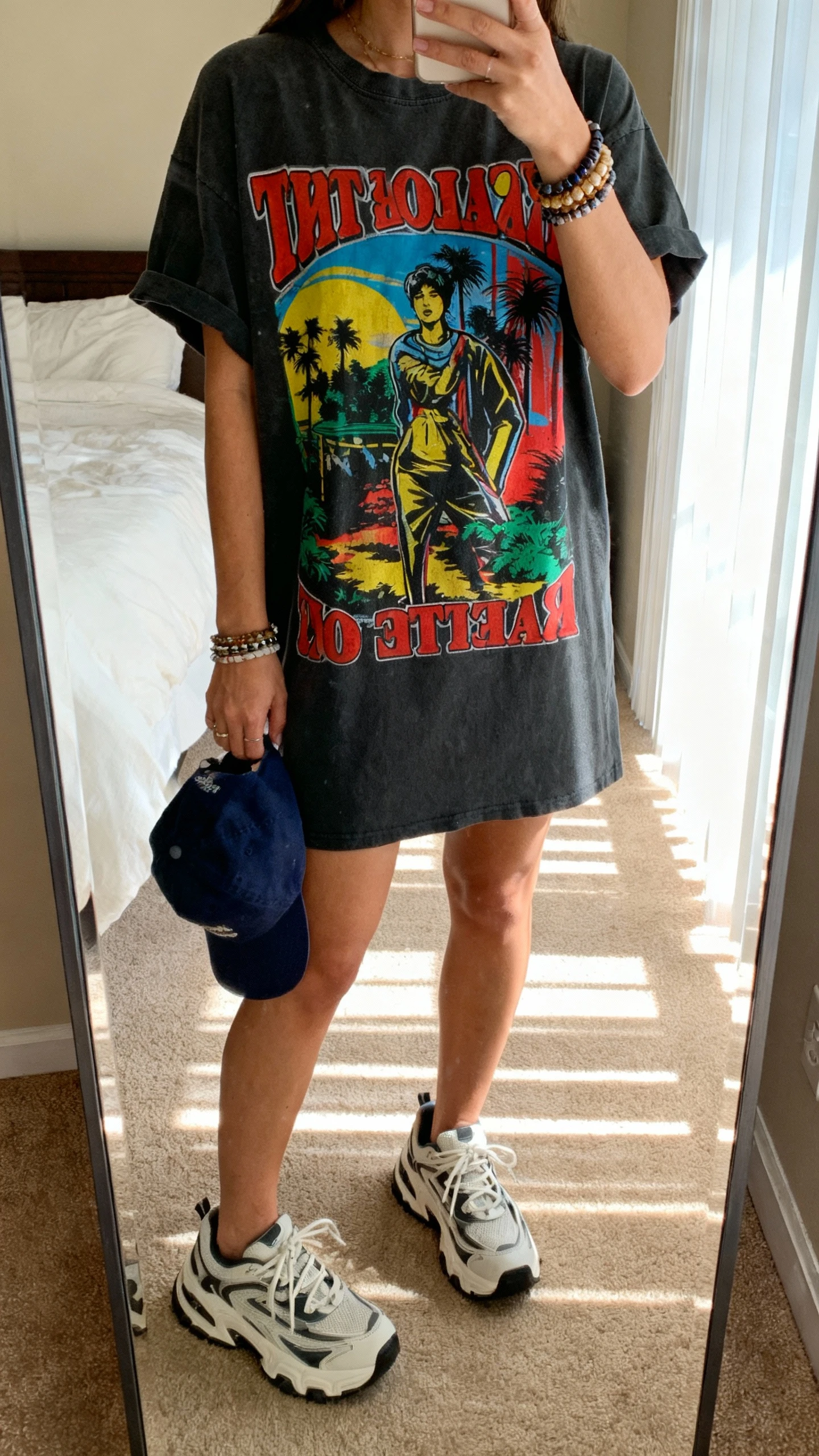 iPhone mirror selfie of a woman in a T-shirt dress with big energy: bold color or graphic, chunky sneakers, stacked bracelets, baseball cap in hand, face not visible, bedroom full-length mirror, bright morning window light, natural iPhone photo.