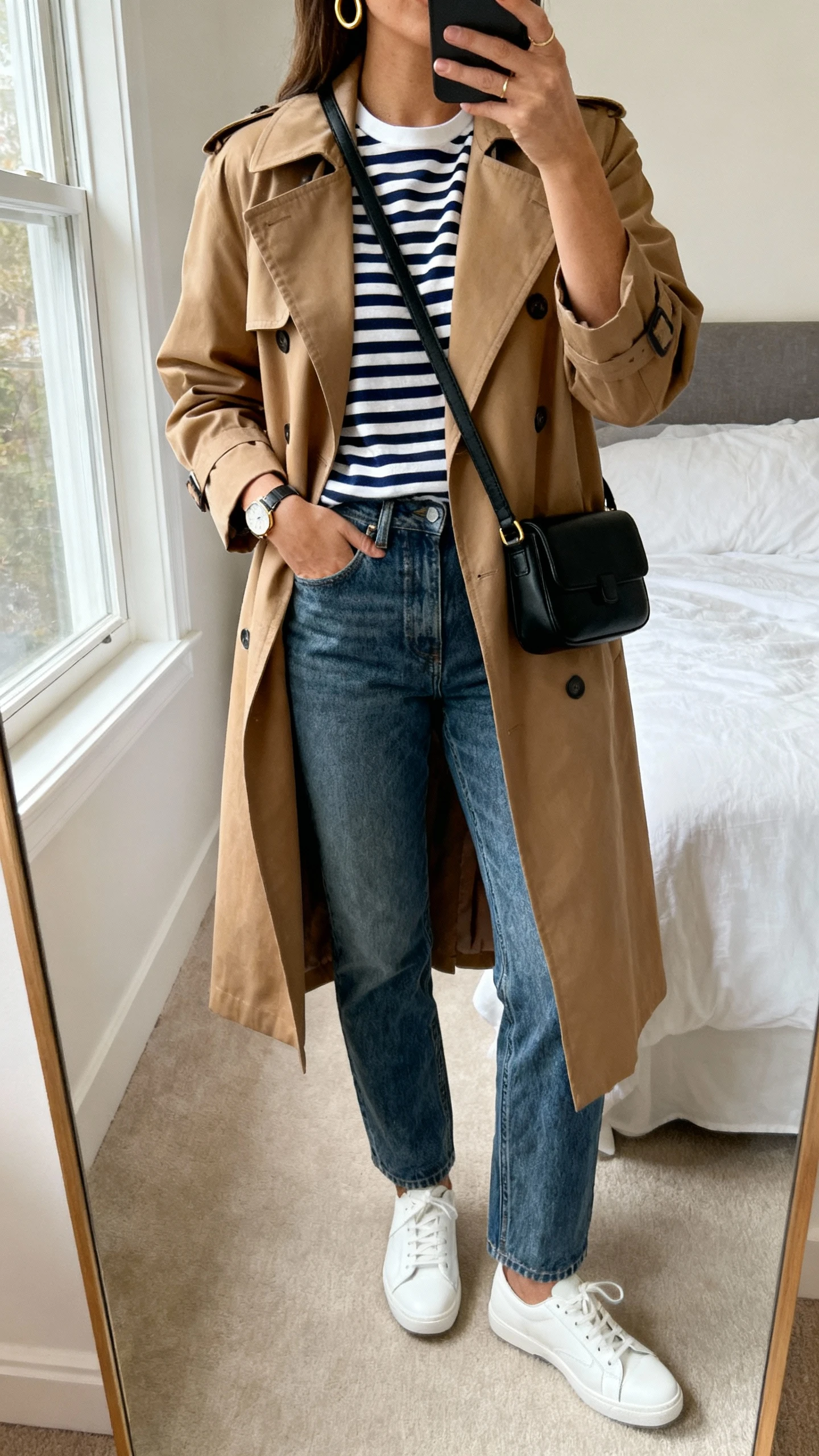 iPhone mirror selfie of a woman in a tan knee-length trench coat over a navy-and-white Breton striped tee, straight-leg medium-wash jeans, white leather sneakers, structured black crossbody, gold hoops and simple watch; bedroom mirror, natural window light, face not visible, cropped at neck, natural iPhone photo quality.