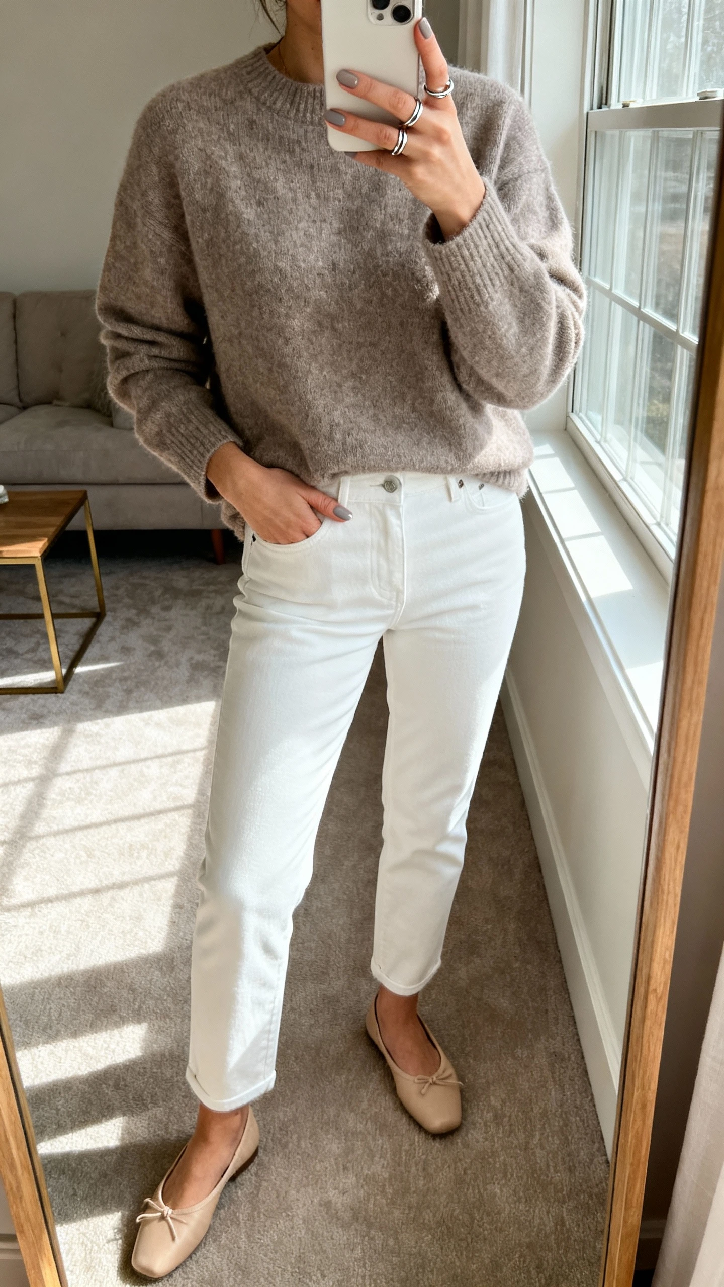 iPhone mirror selfie of a woman in a taupe-gray cashmere sweater, white tailored jeans, beige ballet flats, minimalist silver ring set, soft taupe-gray nails on the phone, face not visible, living room mirror near a window, soft morning light, casual iPhone aesthetic