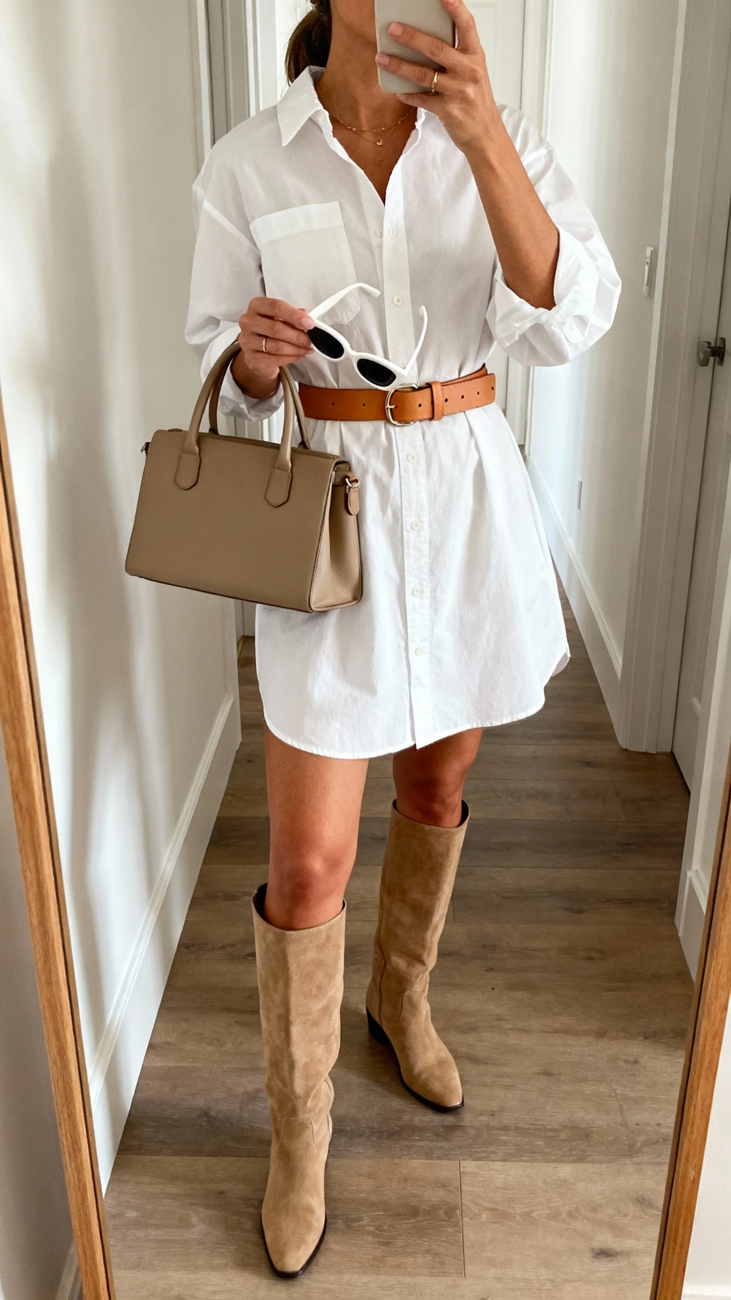 iPhone mirror selfie of a woman in a white cotton poplin shirt dress cinched with a tan leather belt, knee-high boots, top-handle bag, clean-frame sunglasses in hand; hallway mirror, soft indoor light, face not visible, cropped at neck, natural iPhone photo quality.