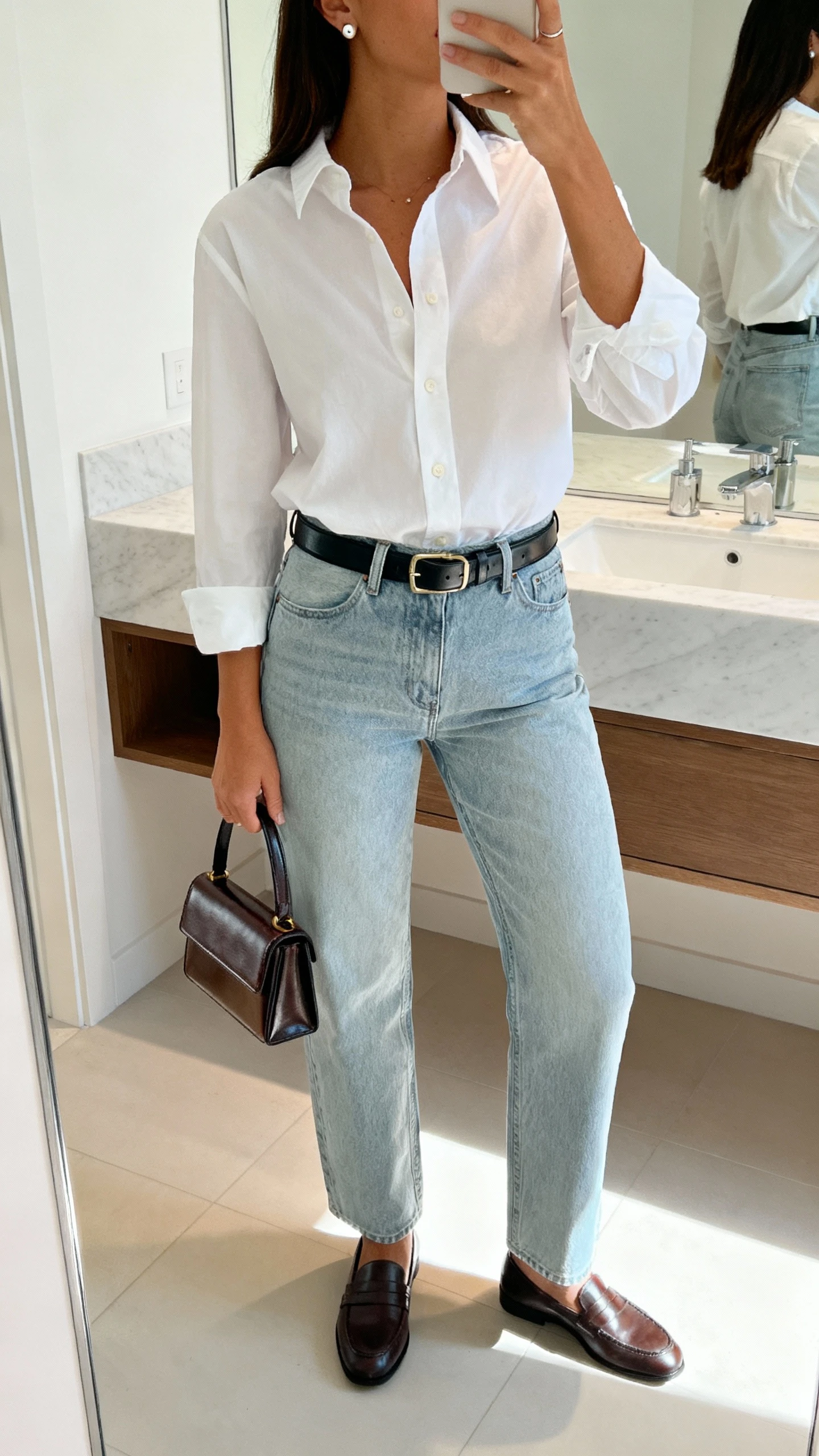 iPhone mirror selfie of a woman in a white cotton button-up tucked into relaxed straight light-wash jeans, loafers, sleek leather belt, structured mini tote, simple stud earrings; bathroom mirror, bright indoor light, face not visible, cropped at shoulders, natural iPhone photo quality.