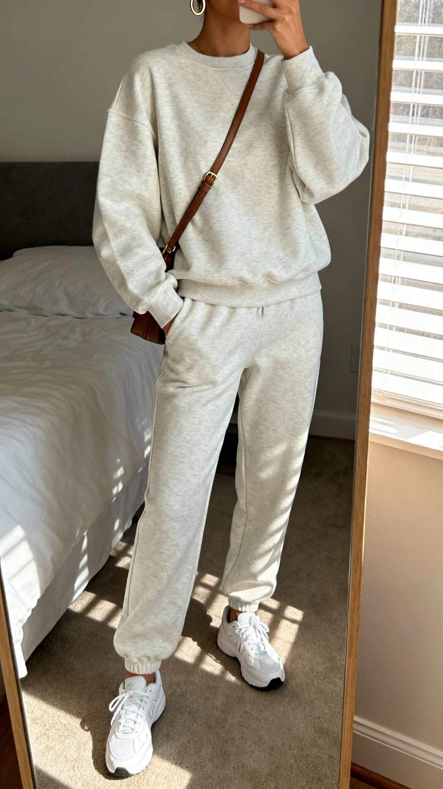 iPhone mirror selfie of a woman in an elevated matching sweatsuit set (tailored sweatshirt and joggers), clean white sneakers, simple hoop earrings, crossbody bag, face not visible, cropped at neck, bedroom mirror with morning window light, natural iPhone photo quality.