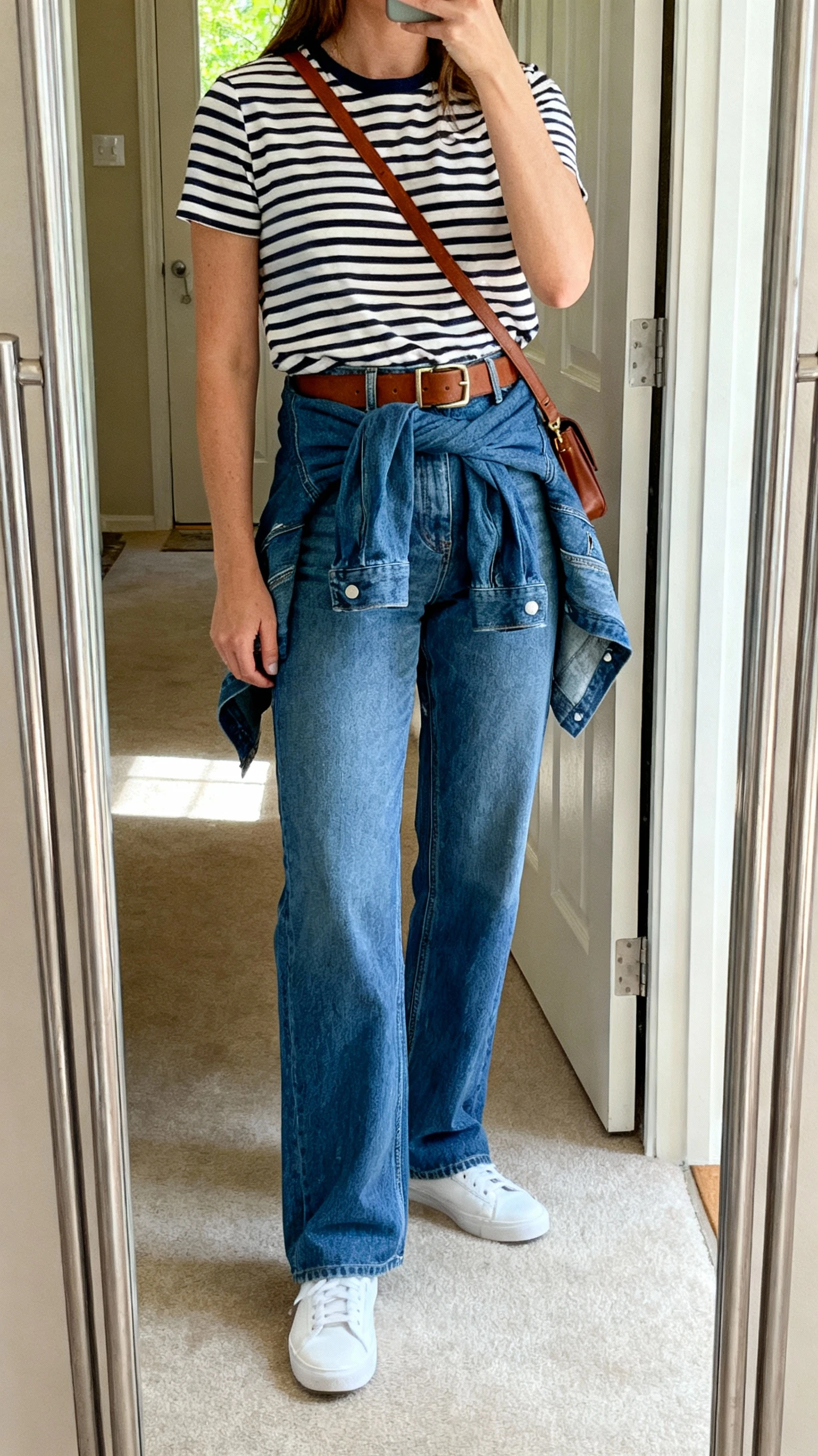 iPhone mirror selfie of a woman in straight-leg blue jeans with a striped Breton tee, white sneakers, brown belt, denim jacket tied around waist, crossbody bag; face not visible, full-length mirror in entryway, natural daylight from doorway, casual iPhone shot.