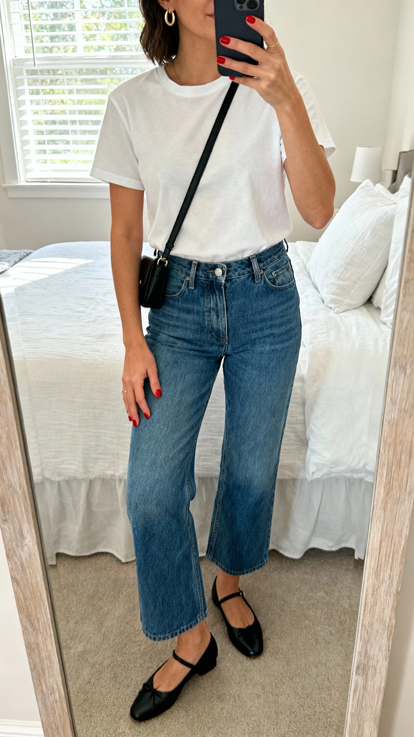 iPhone mirror selfie of a woman showing short cherry red nails with a simple white tee, high-rise blue jeans, black ballet flats, small gold hoops, and a black crossbody, face not visible, cropped at neck, bedroom mirror, natural window light, natural iPhone photo quality.