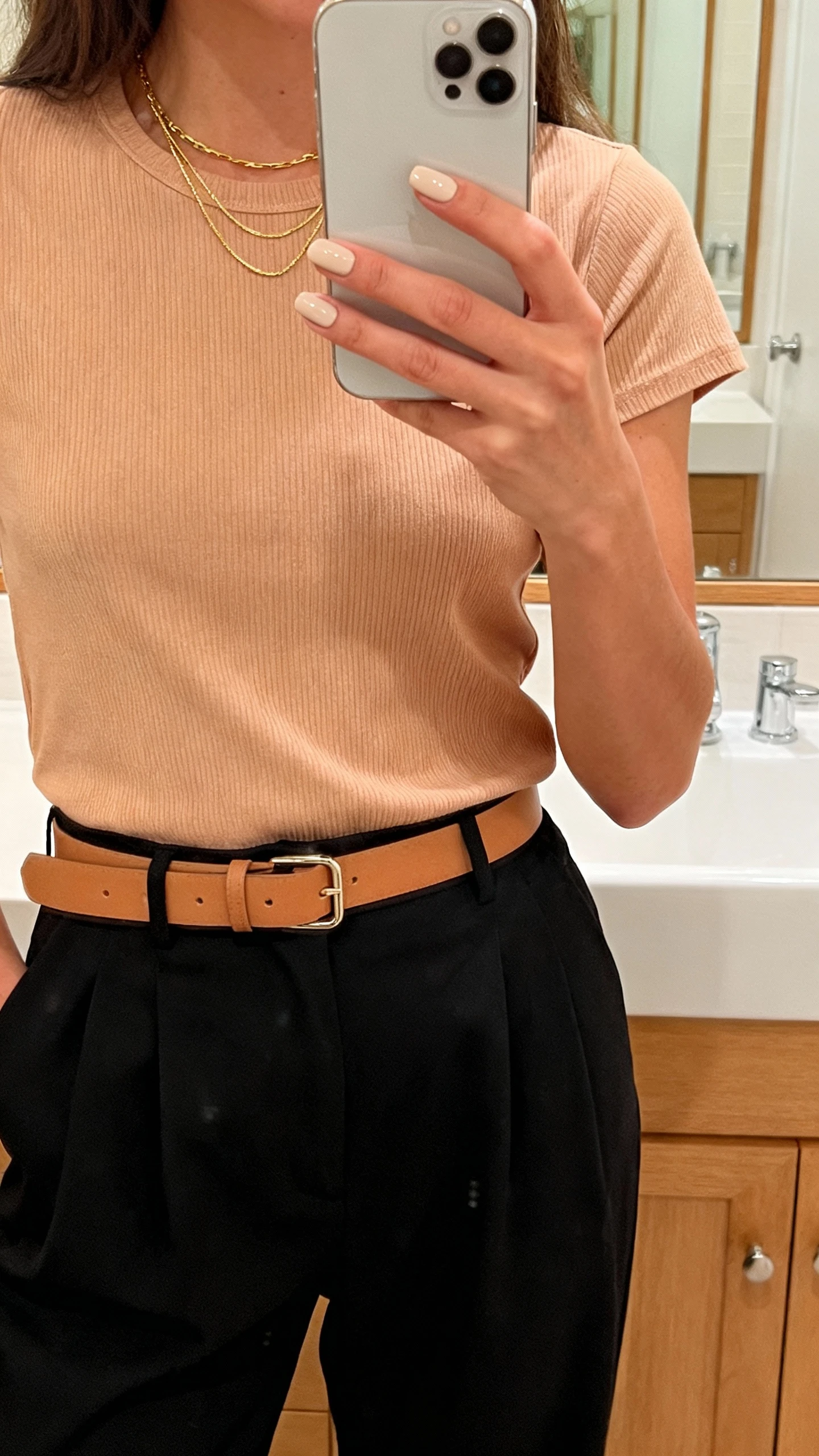 iPhone mirror selfie of a woman wearing a beige-peach ribbed tee, high-waisted black trousers, tan belt, delicate layered gold necklaces, soft beige-peach nails holding her phone, face not visible, bathroom mirror, warm indoor lighting, natural iPhone aesthetic