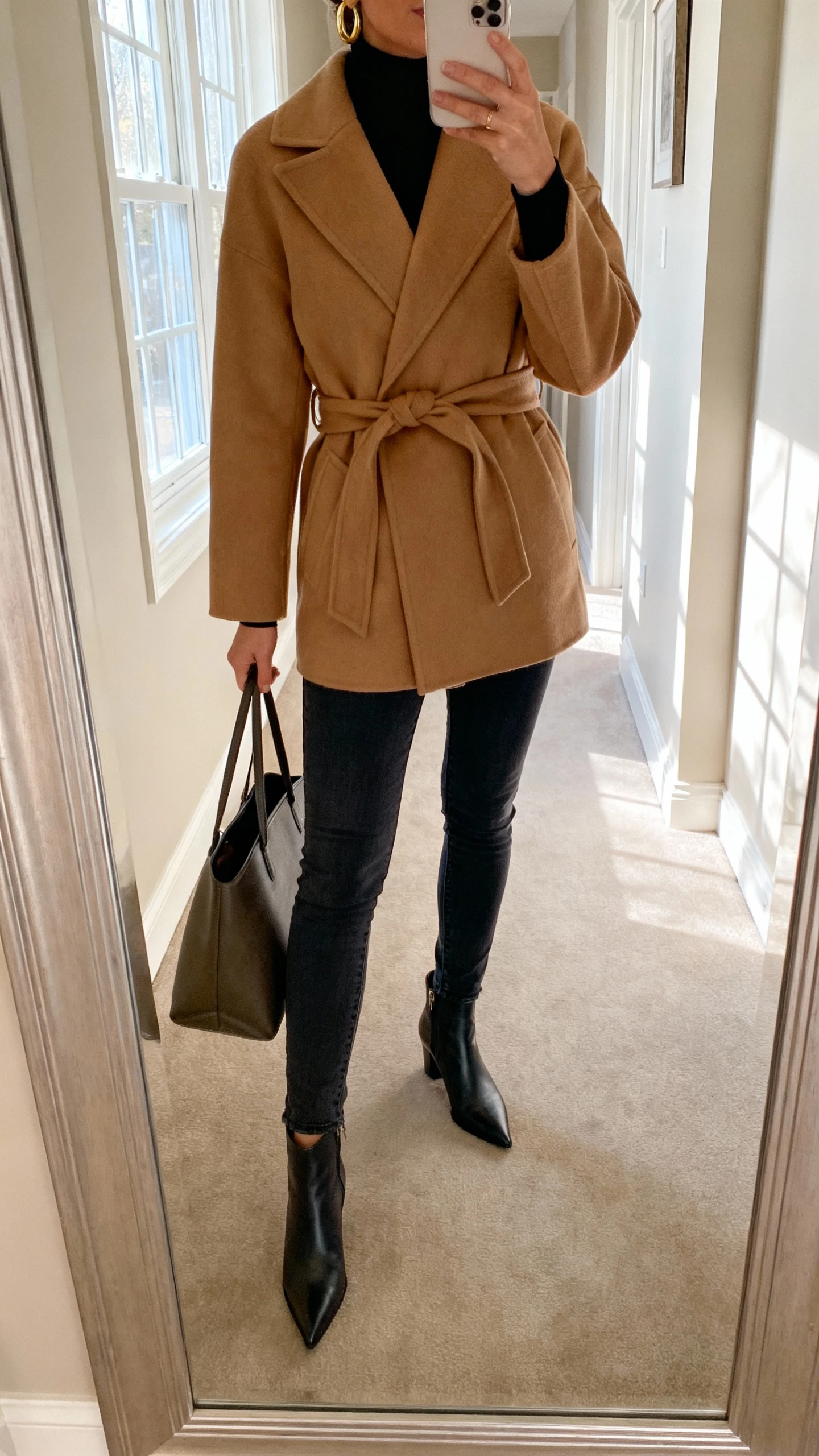 iPhone mirror selfie of a woman wearing a camel belted wrap coat over a black turtleneck, slim dark jeans, pointed ankle boots, structured tote, gold hoop earrings; face not visible, cropped at neck, hallway mirror after errands, soft afternoon window light, natural iPhone photo quality.