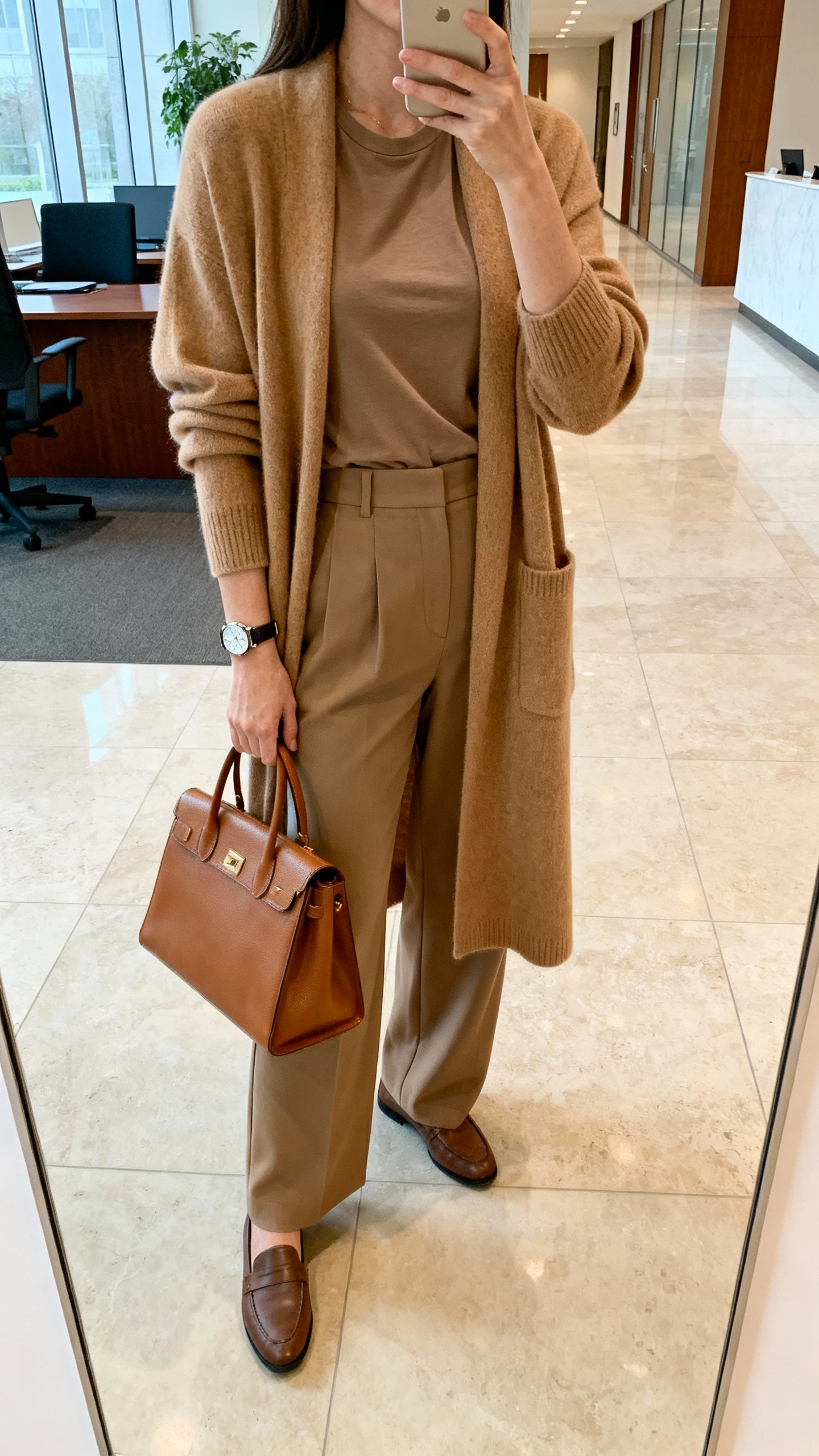 iPhone mirror selfie of a woman wearing a cardigan-coat in camel over a fitted tee, high-waist wide-leg trousers, loafers, structured leather tote, watch, face not visible, cropped at neck, office lobby mirror with soft indoor lighting, natural iPhone photo quality.