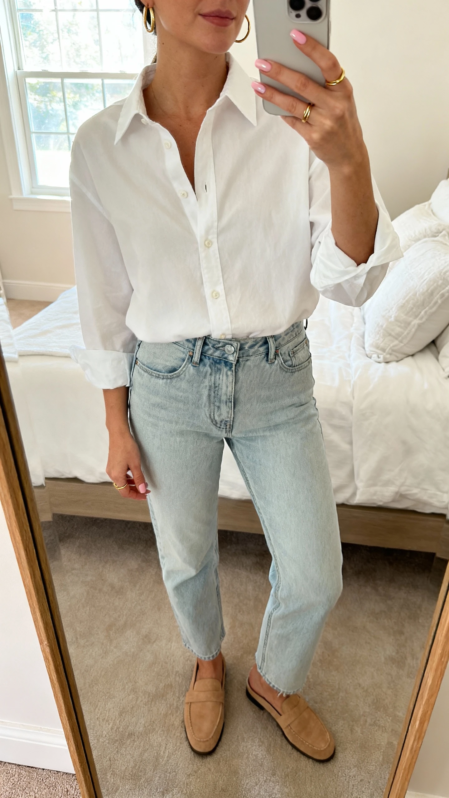 iPhone mirror selfie of a woman wearing a crisp white button-up tucked into light-wash straight jeans, tan loafers, slim gold hoop earrings, dainty gold rings showcasing classic sheer pink nails, face not visible, cropped at neck, bedroom mirror with soft morning window light, natural iPhone photo quality