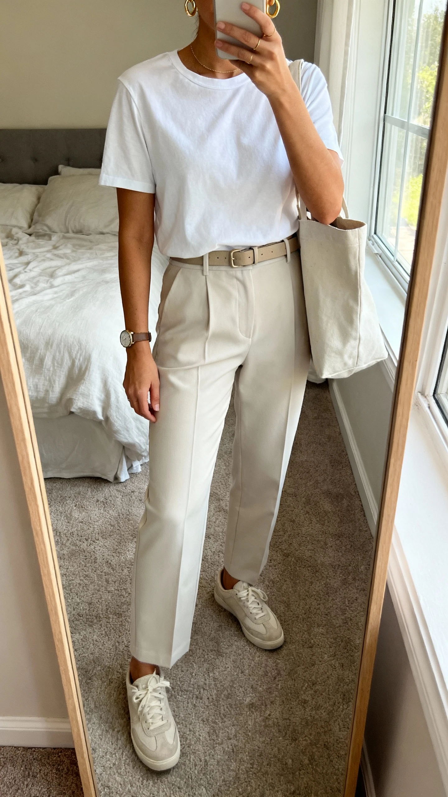 iPhone mirror selfie of a woman wearing a crisp white elevated tee tucked into tailored trousers, minimalist sneakers, slim belt, delicate gold hoops and watch, tote bag on arm, face not visible, cropped at neck, bedroom mirror with morning window light, natural iPhone photo quality.
