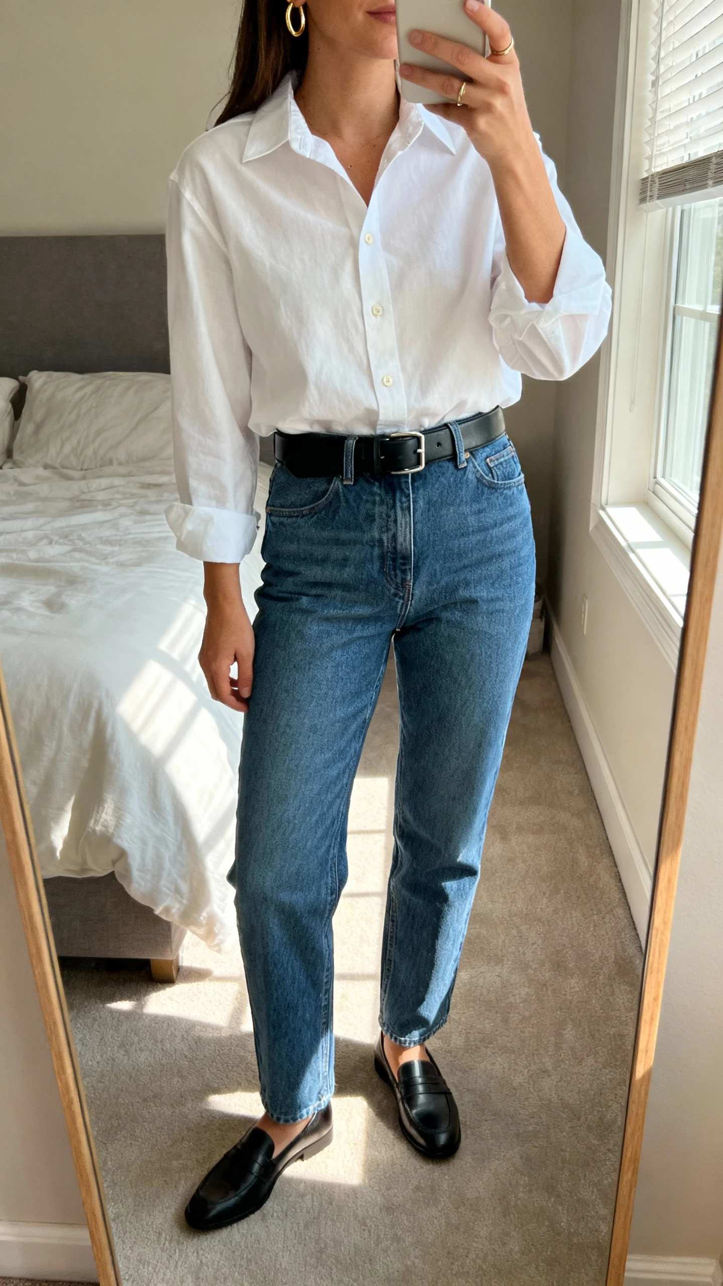 iPhone mirror selfie of a woman wearing a crisp white button-down tucked into straight-leg blue jeans, black leather belt, loafers, simple gold hoops, face not visible, cropped at neck, bedroom mirror with morning window light, natural iPhone photo quality.